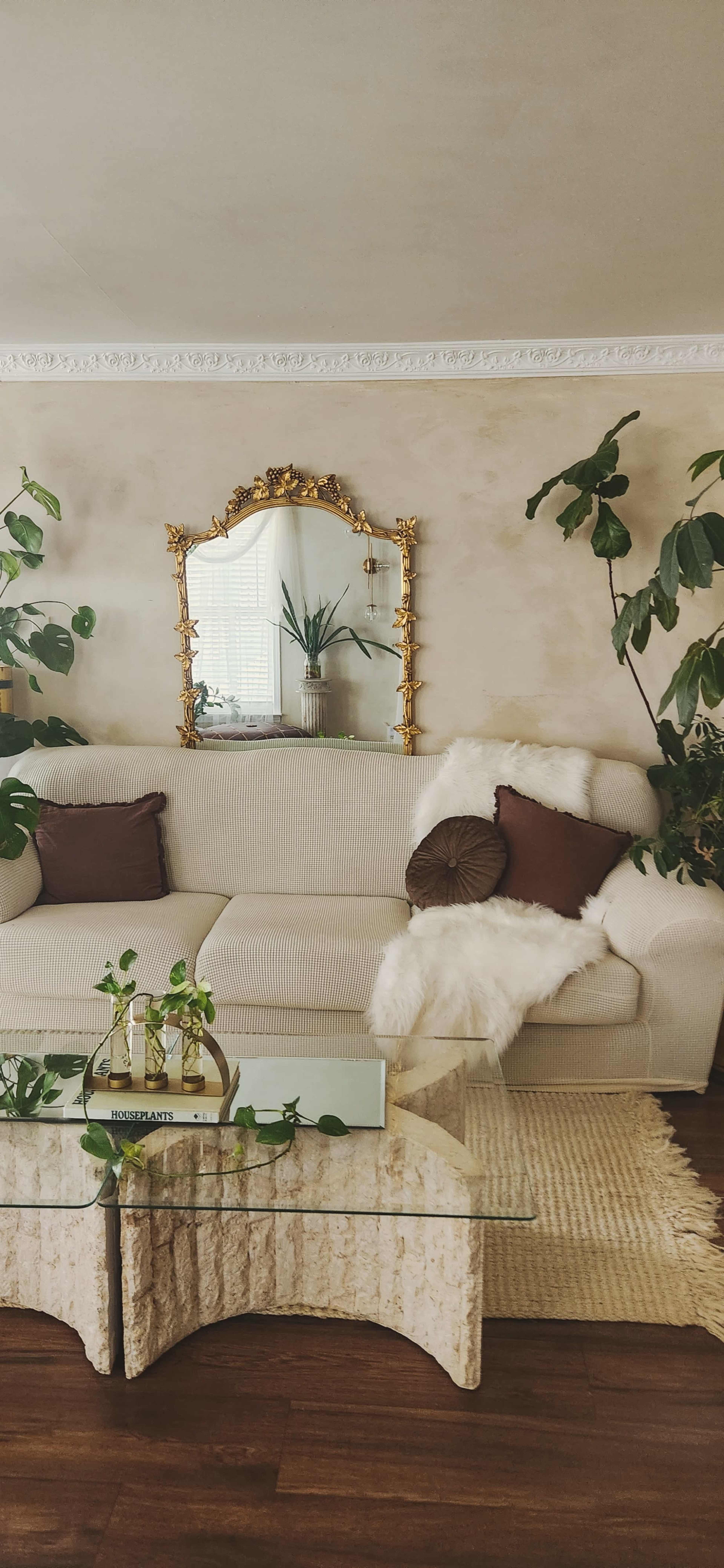 The image features a living room with a white sofa adorned with decorative pillows, a glass coffee table, and various indoor plants alongside a vintage mirror on the wall.