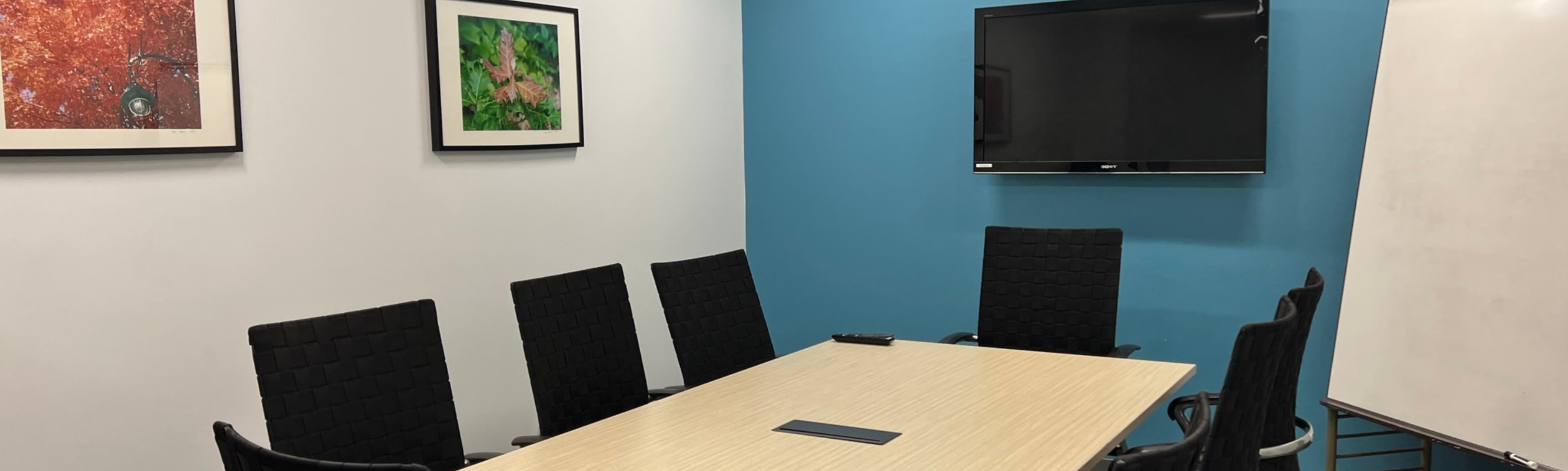 A conference room features a wooden table surrounded by six black chairs, with framed artwork on the walls and a television mounted on a blue wall.