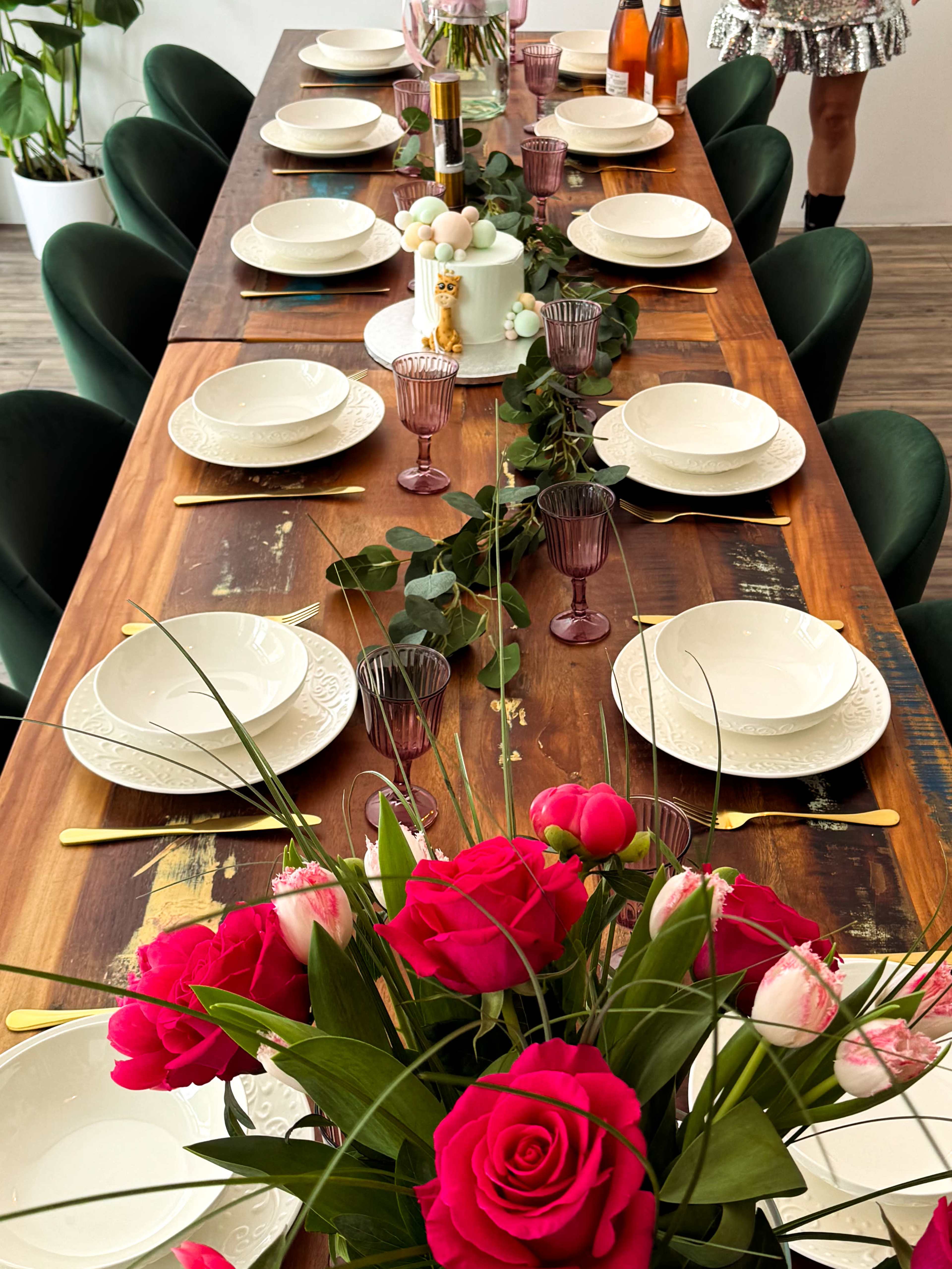 A long wooden dining table is elegantly set with plates, glasses, and a floral centerpiece featuring pink roses.