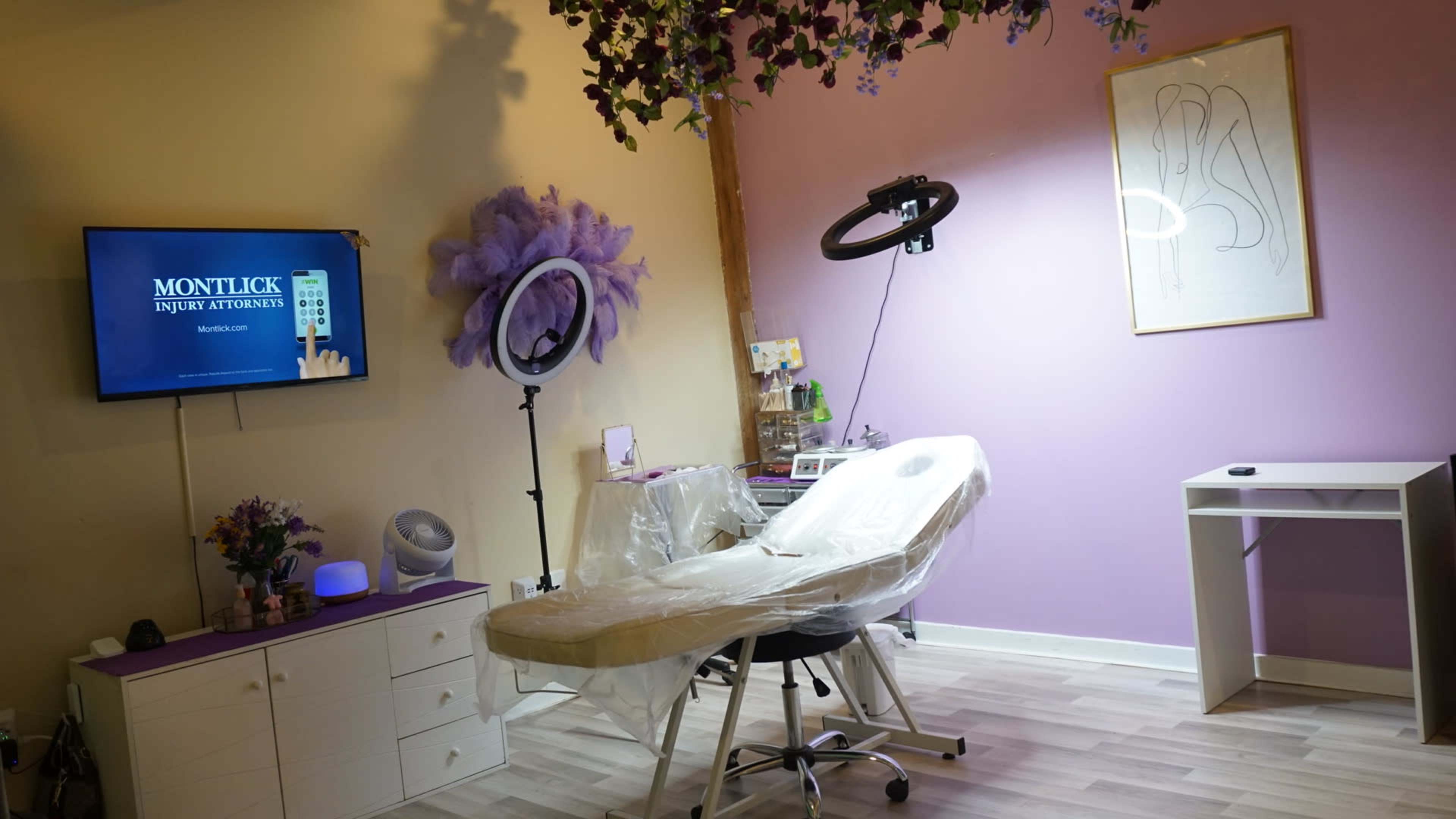The image shows a beauty treatment room featuring a treatment chair, a makeup mirror with lighting, a television on the wall, and decorative elements including flowers and a framed artwork.