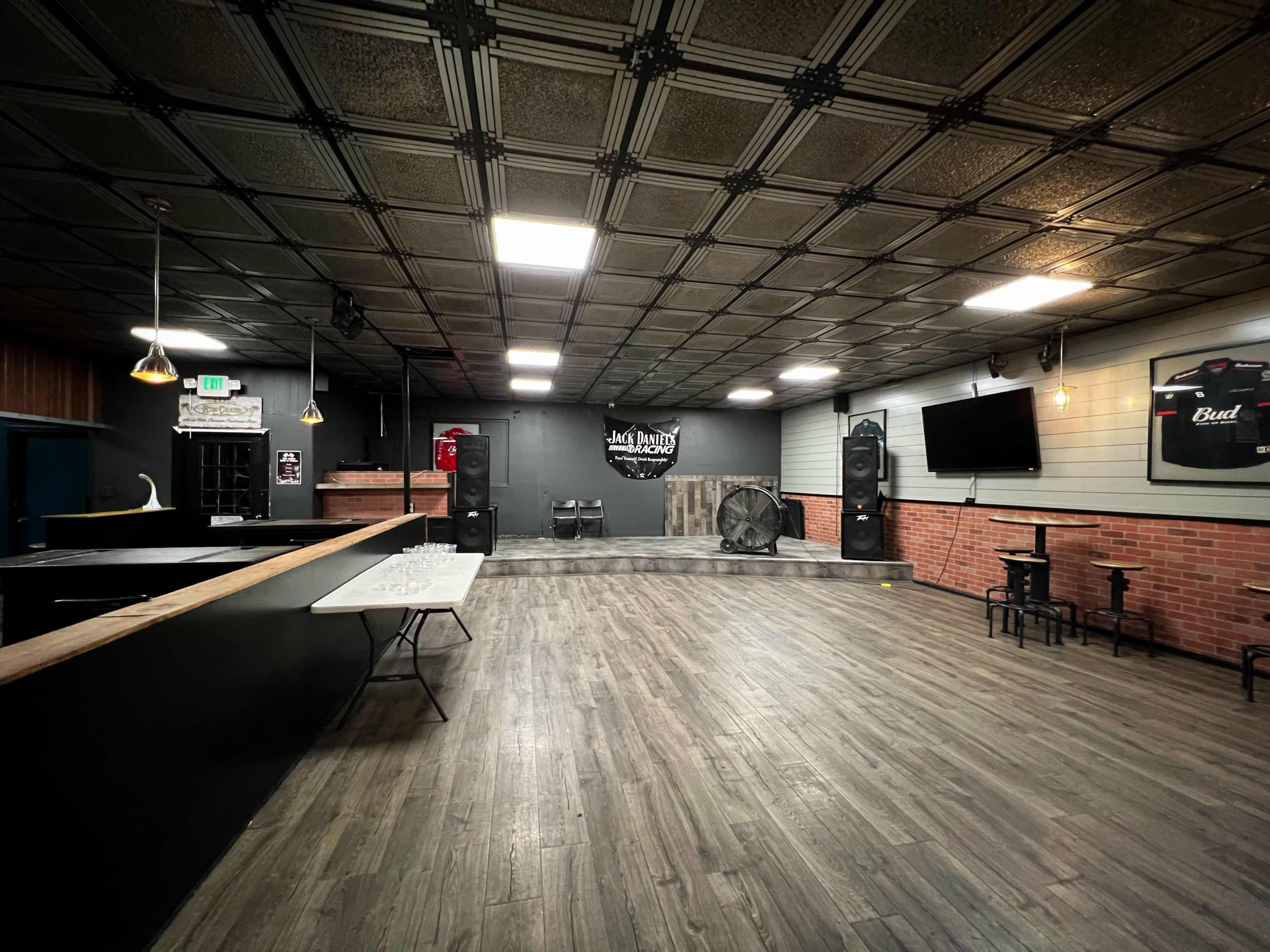 An empty performance space with a wooden floor, a bar area, and a stage equipped with speakers and a large fan at one end.