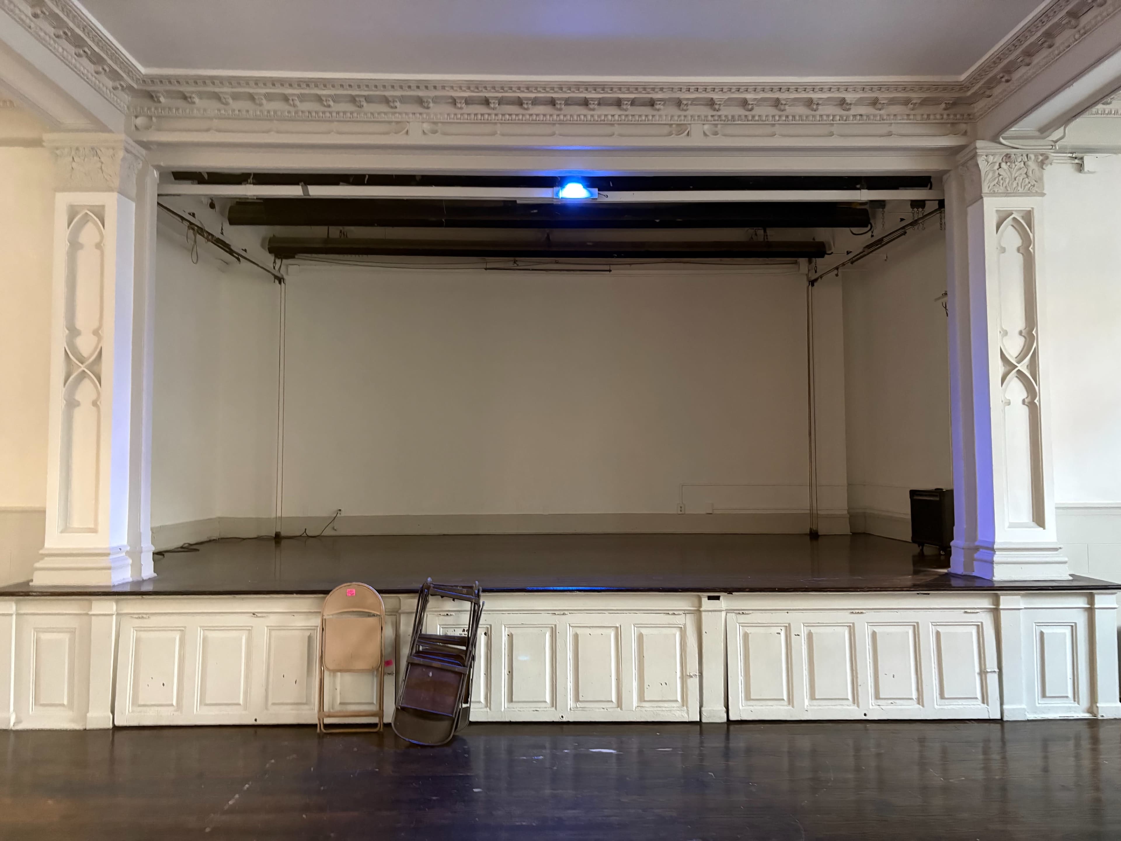 The image shows a vacant stage with a simple wooden floor and a white backdrop, flanked by an empty chair near the front.