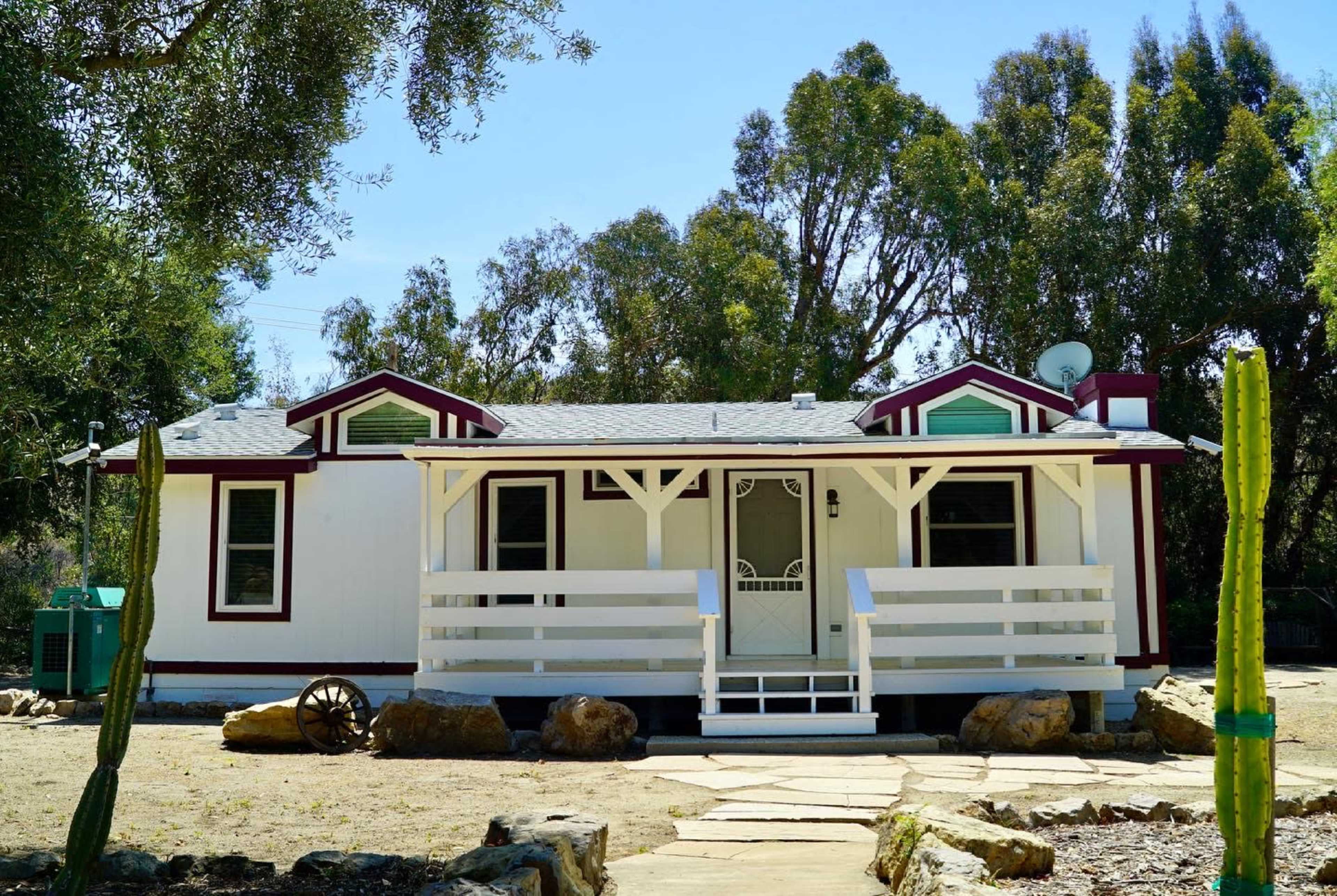 A white house with a front porch, surrounded by trees and cacti, is set in a rural landscape.