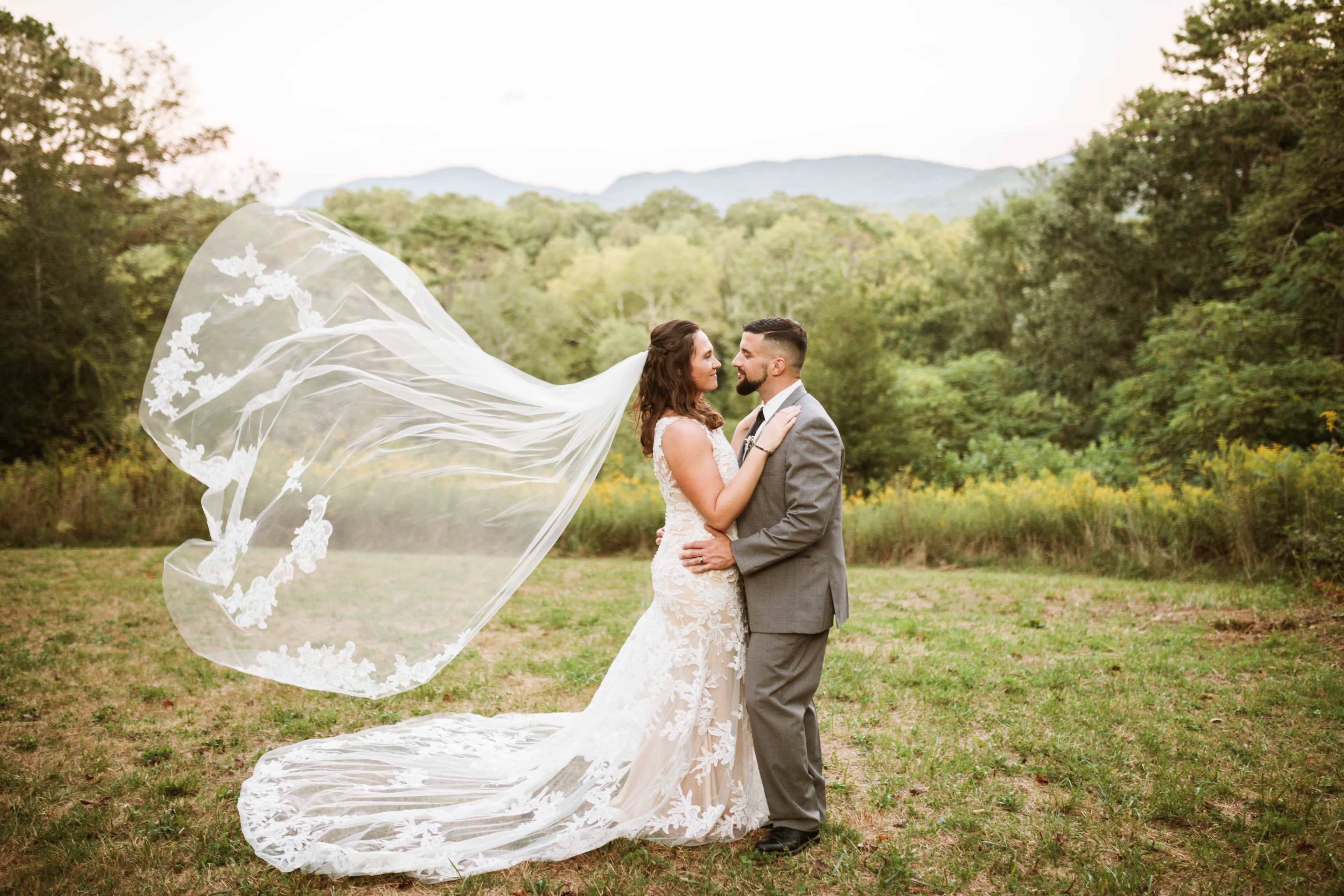A bride and groom embrace in a grassy field with mountains in the background, as the bride's veil flows in the breeze.