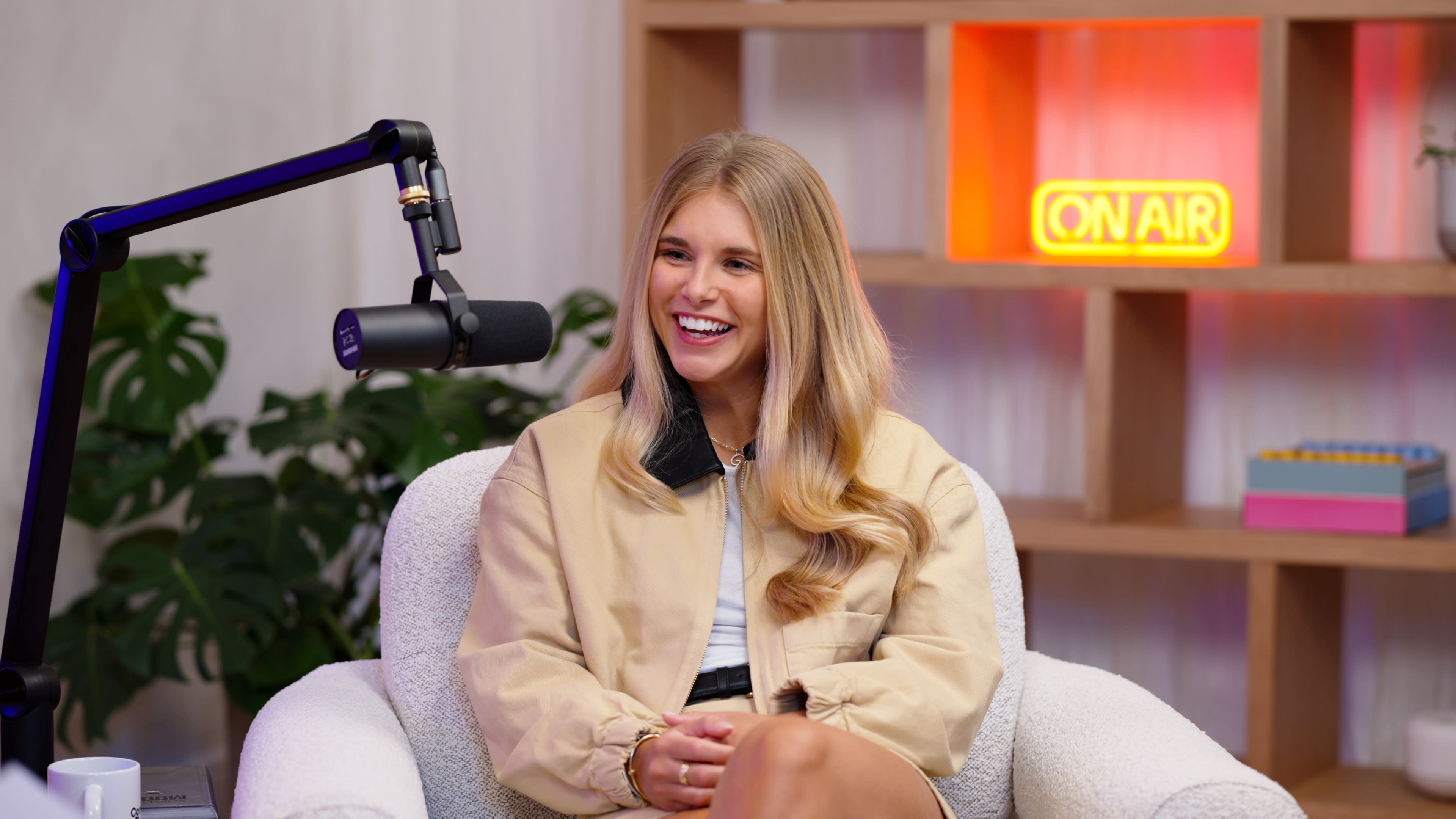 A person with long hair sits in a cozy chair, smiling while speaking into a microphone in a modern studio setting.