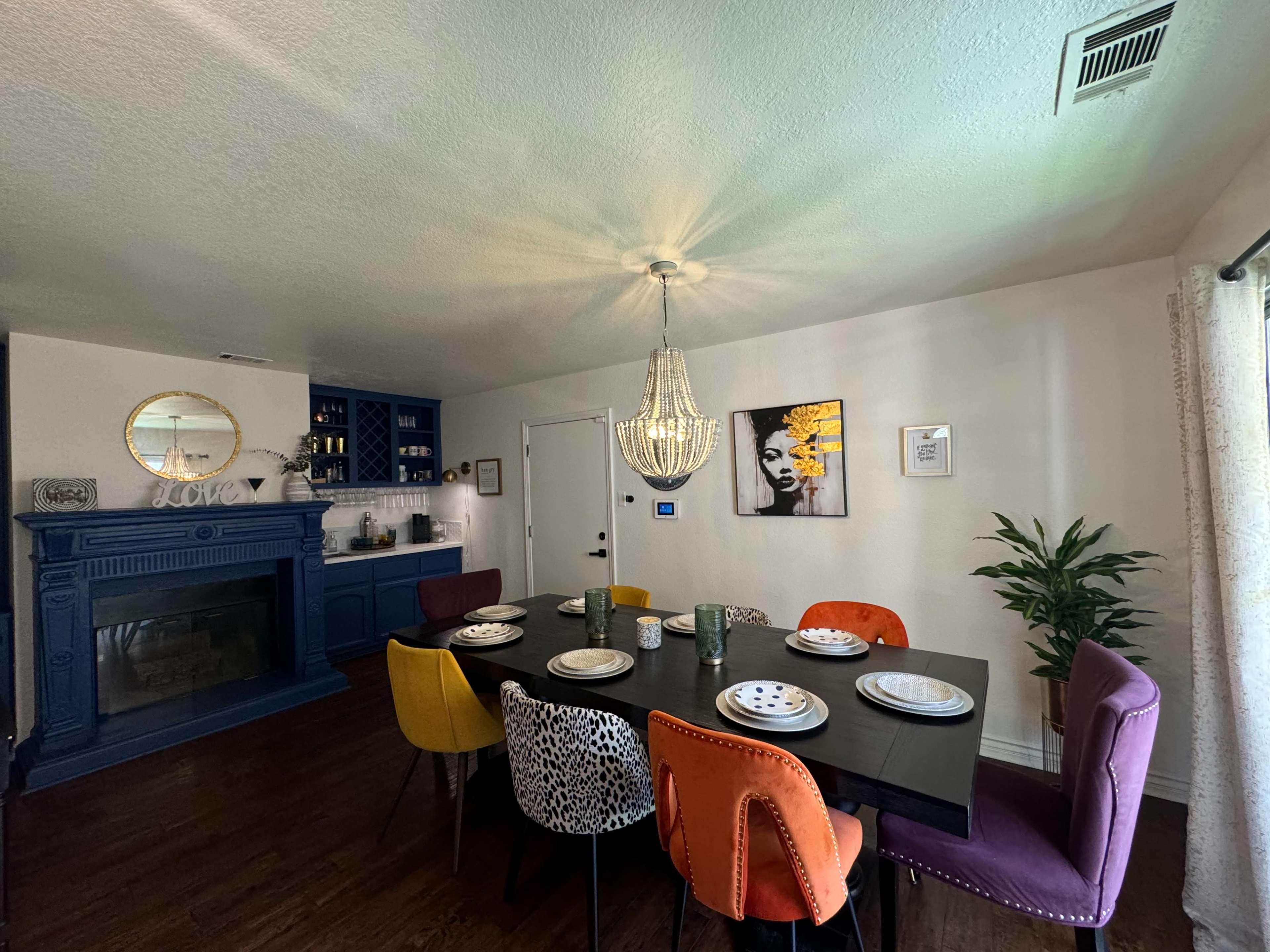 A dining area features a large table set for six with colorful chairs and a chandelier overhead, alongside a wall with artwork and a fireplace.