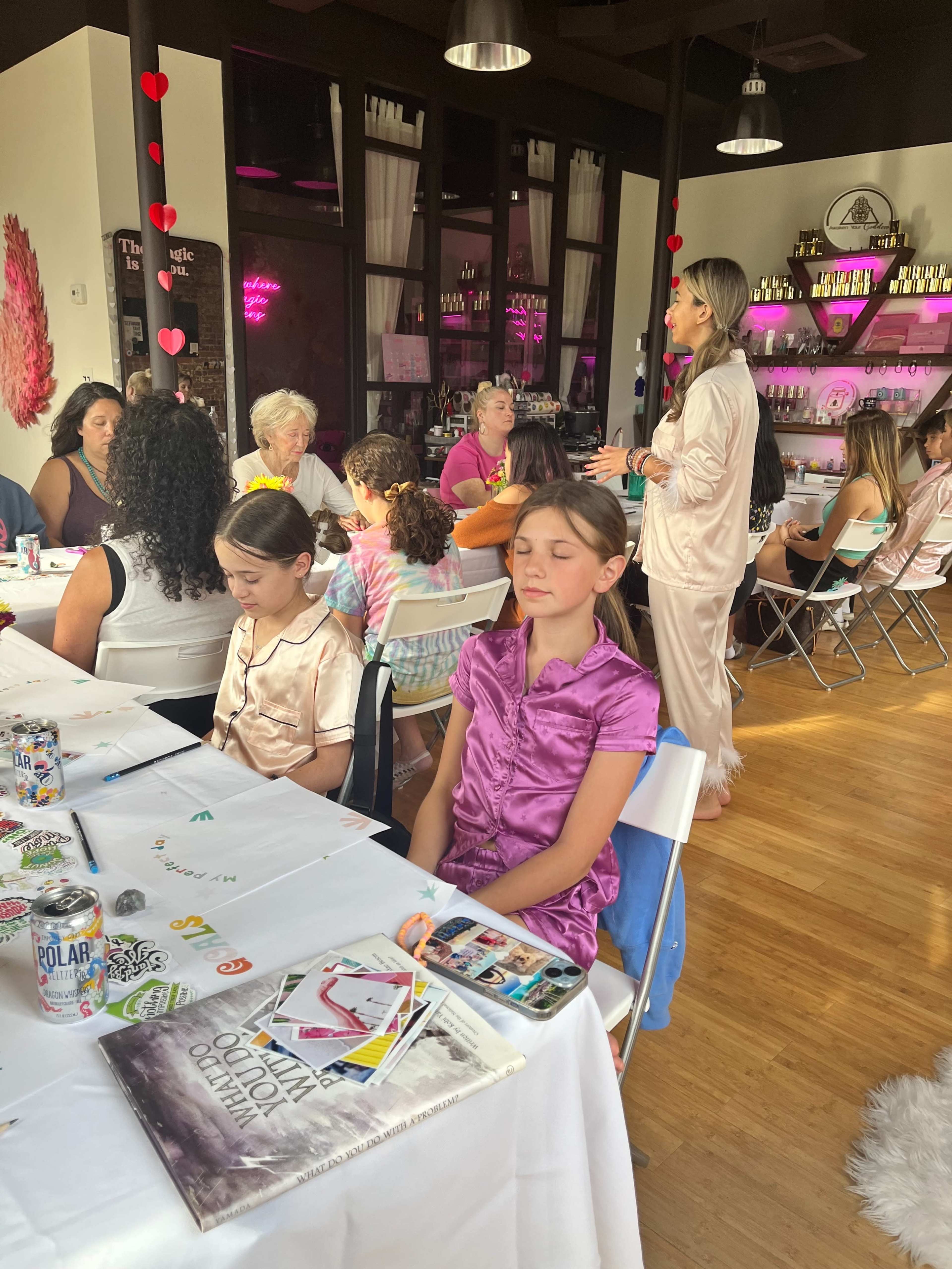 A group of children and adults gather around tables in a brightly lit room, with some participants wearing pajamas and engaging in activities.