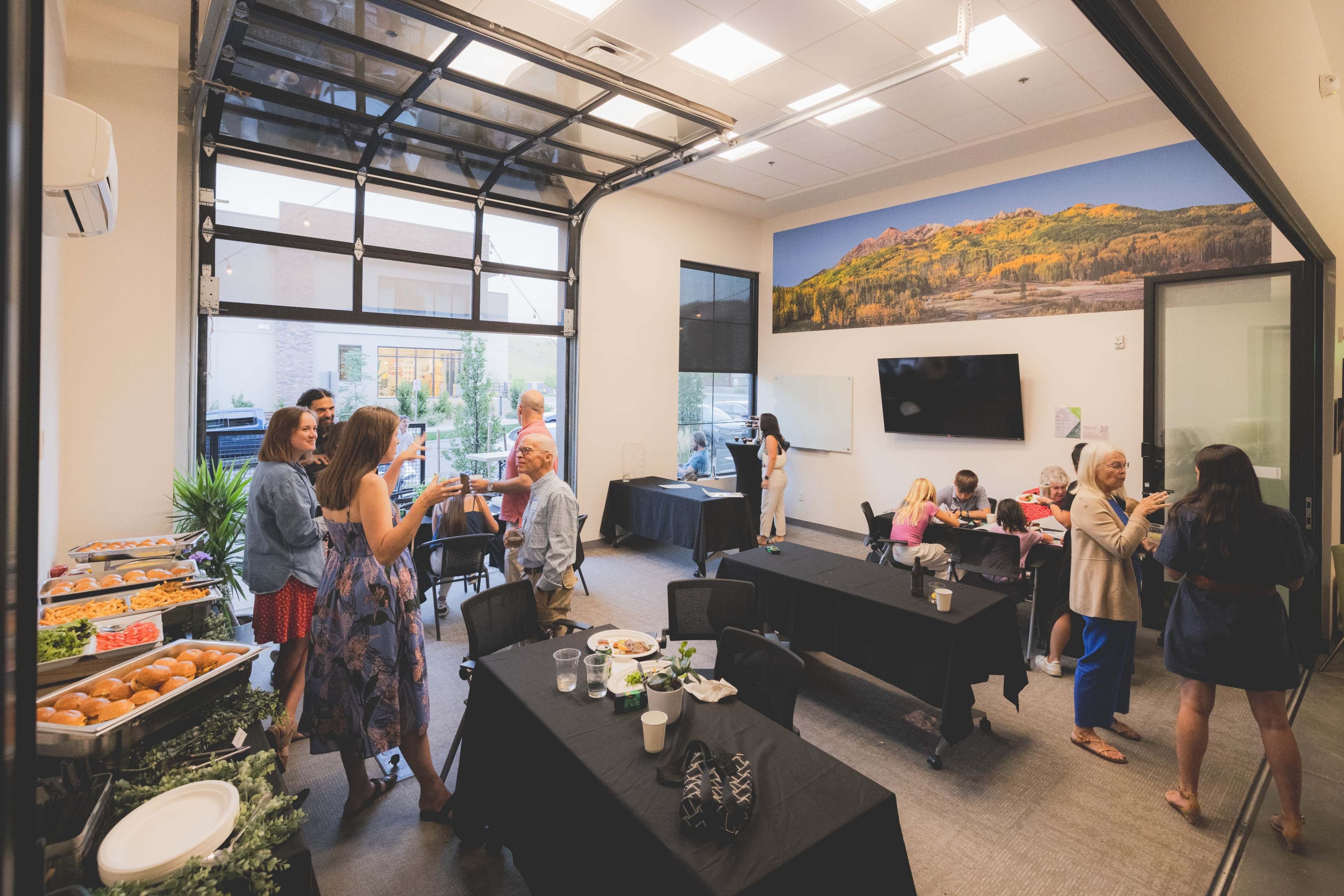 The image shows a modern meeting room with people socializing, tables with food, and a large mural on the wall.
