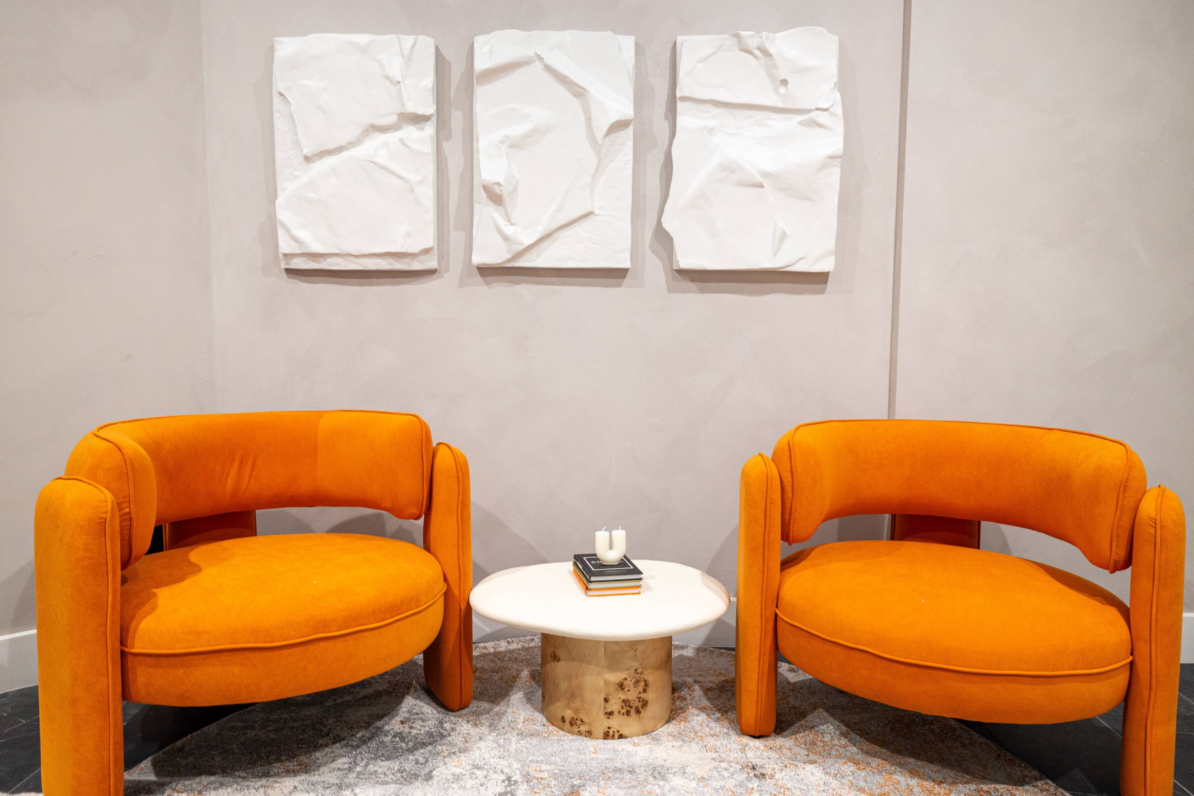 Two orange velvet chairs are positioned facing each other in front of a small round table, with three textured white artworks mounted on the gray wall behind them.