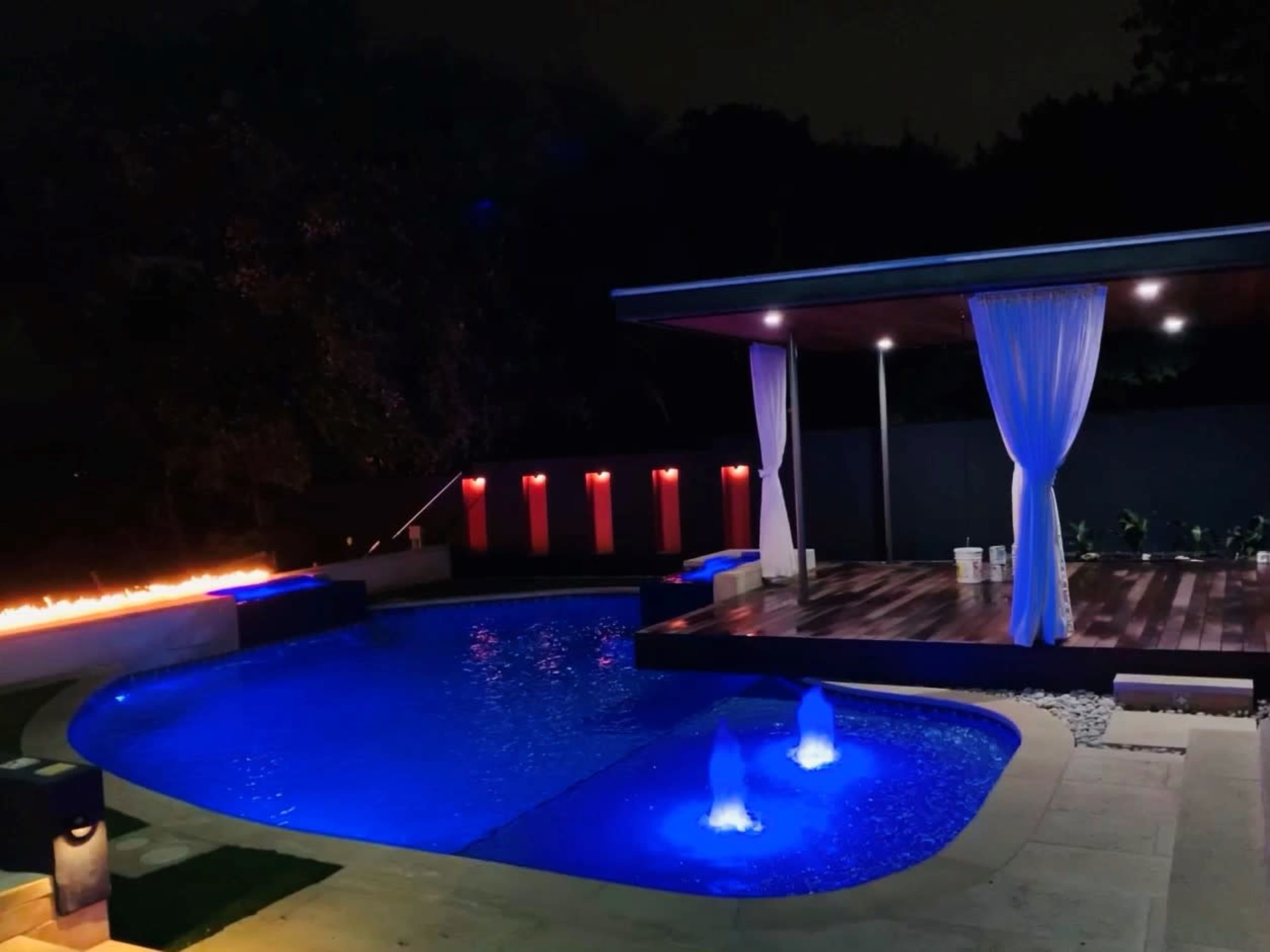 The image shows a modern backyard with a swimming pool featuring illuminated fountains and a covered deck, all set against a night sky.