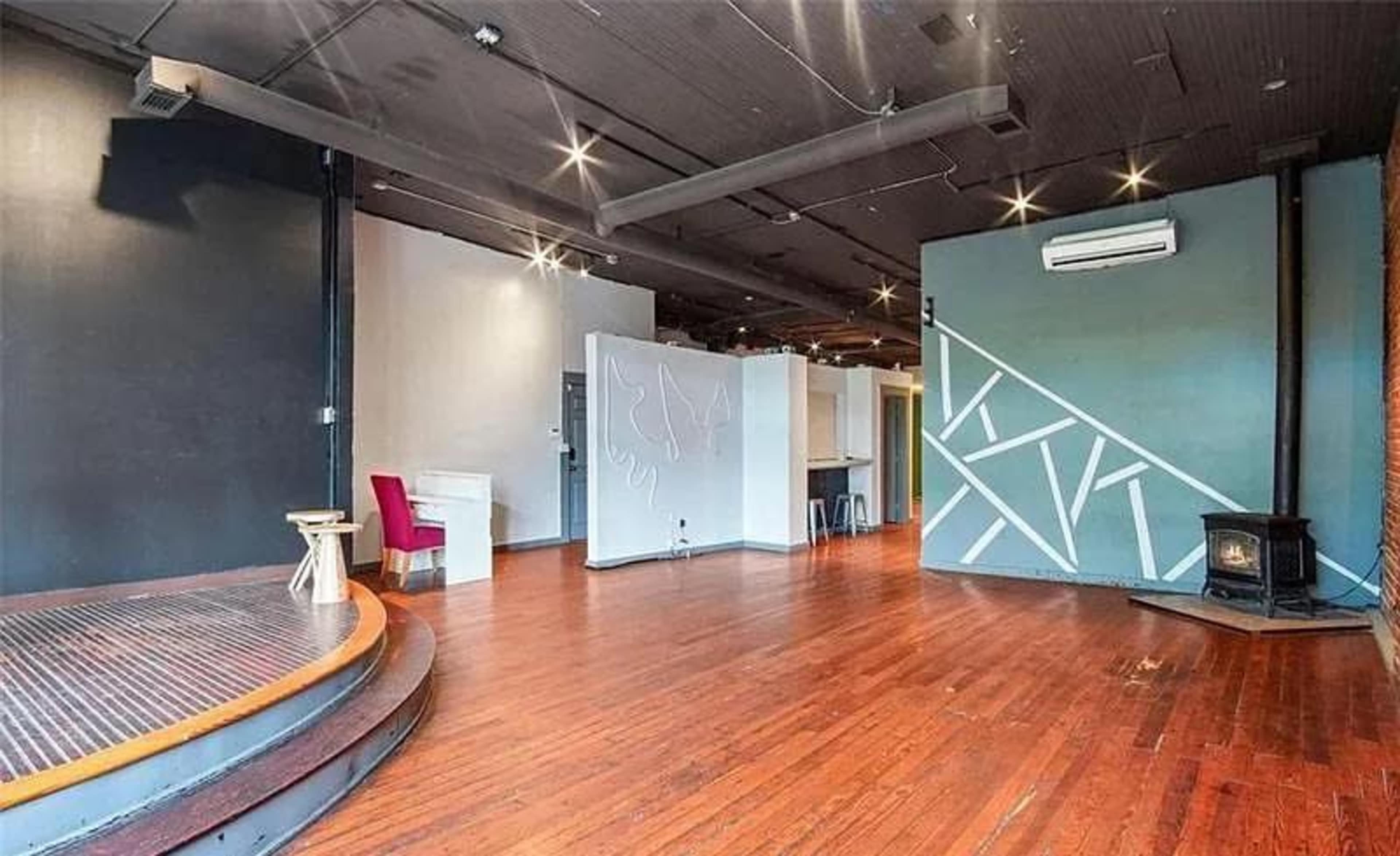 The image shows a spacious, partially furnished room with dark wooden flooring, featuring a wood stove and distinct wall colors.