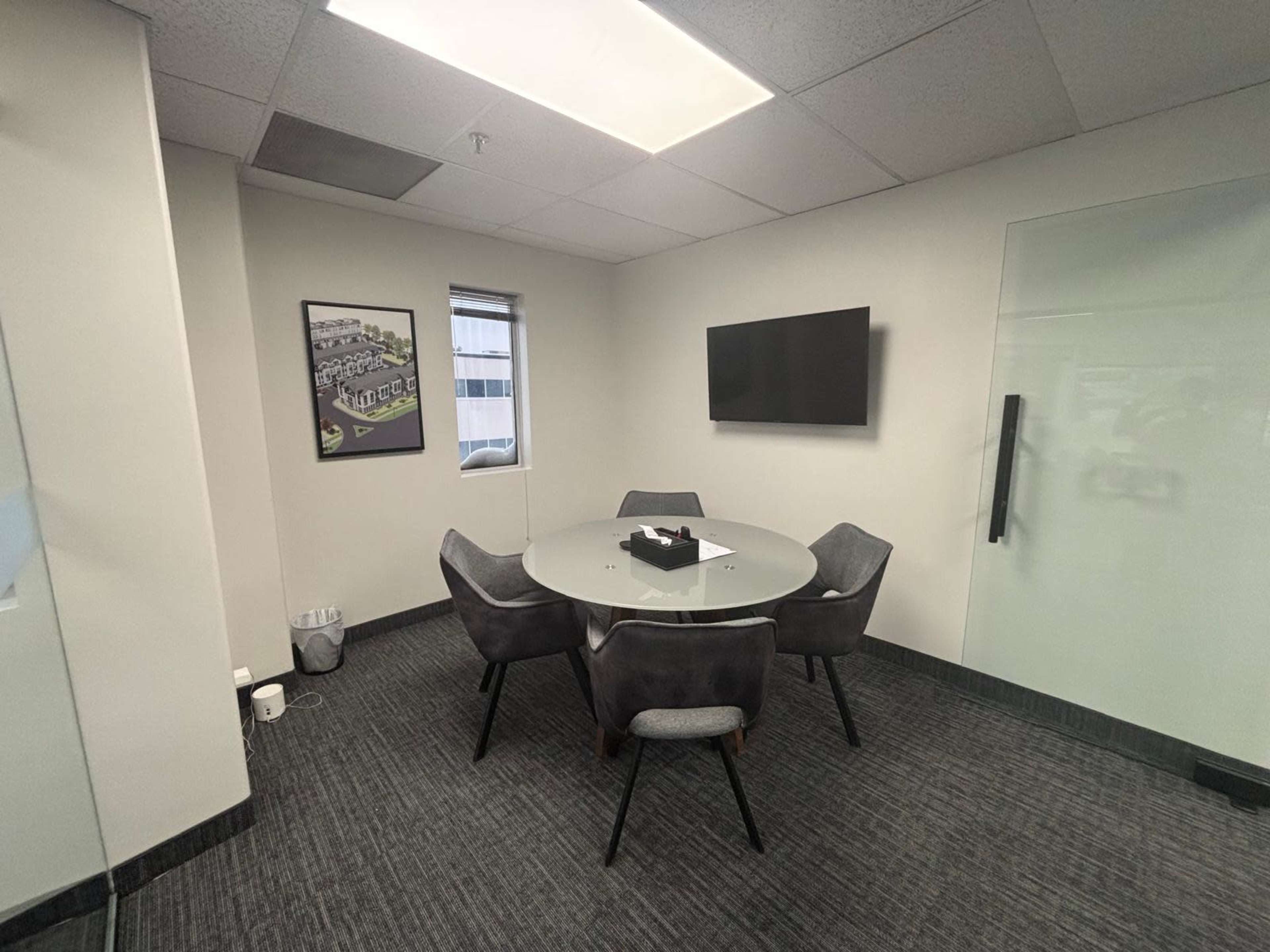 A conference room features a round glass table surrounded by four gray chairs, with a wall-mounted television and a window to the outside.