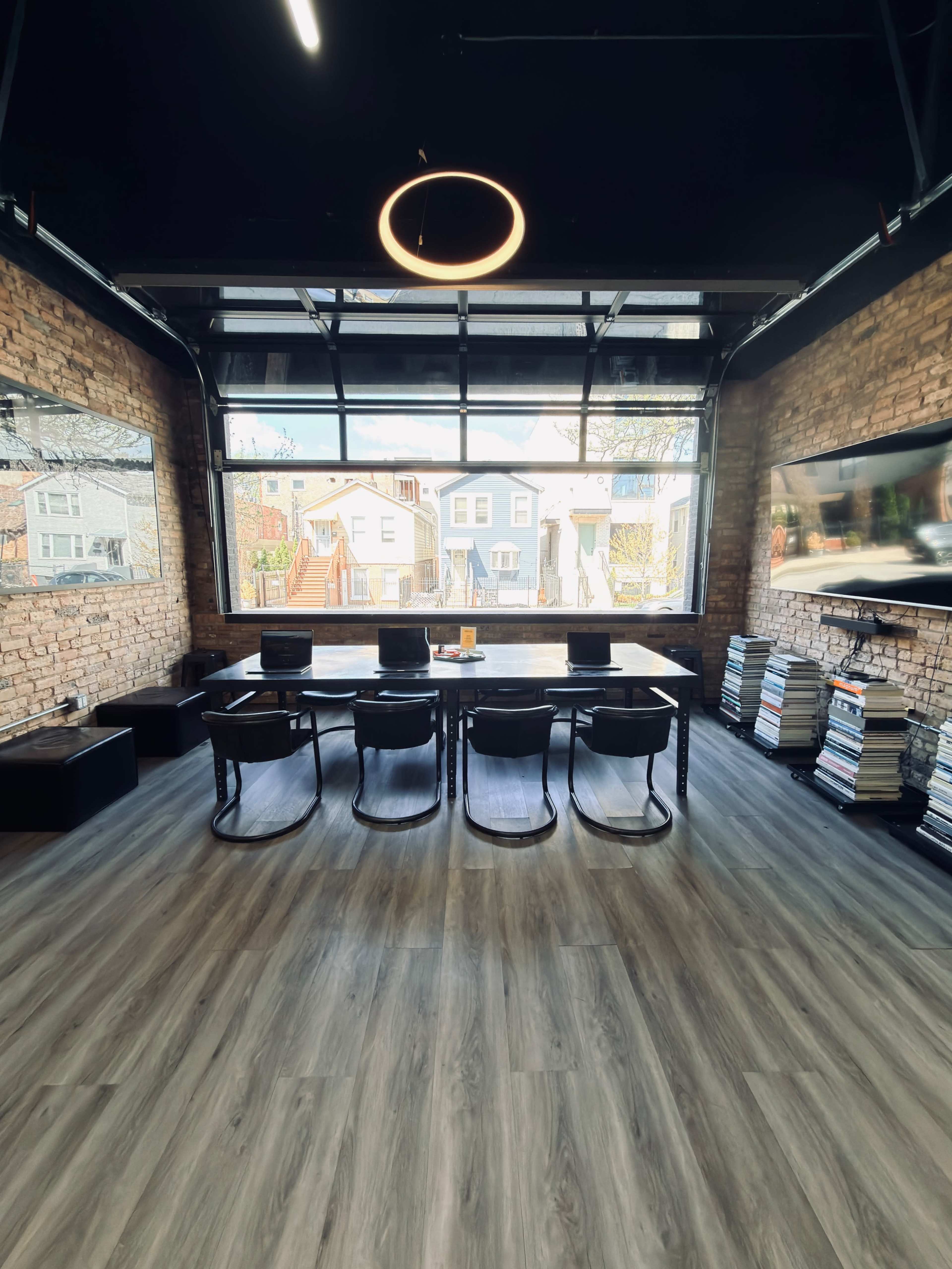 A modern conference room features a large table with black chairs, large windows allowing natural light in, and brick walls adorned with two flat-screen monitors.