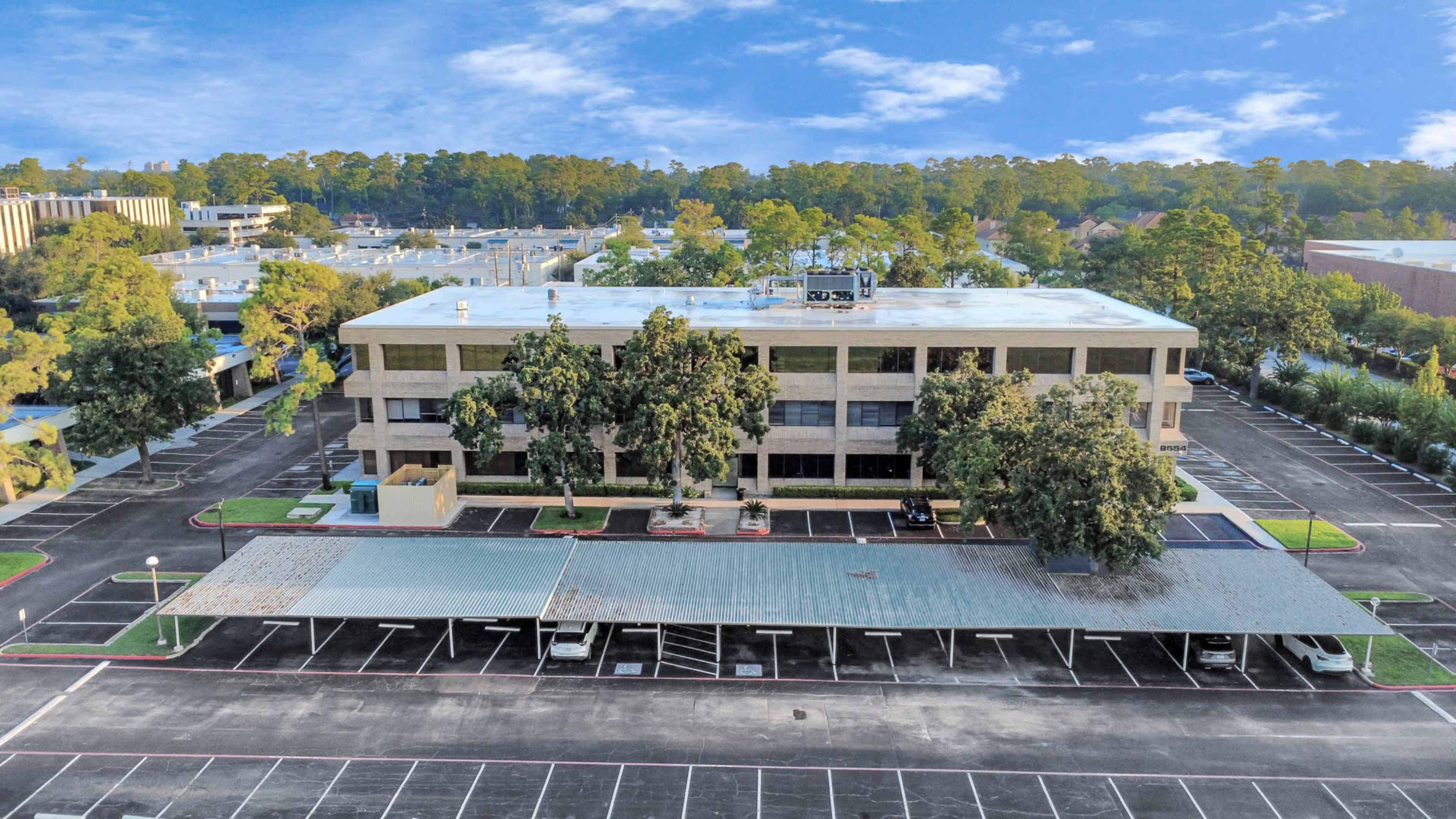 A four-story office building with a parking lot and covered carports is surrounded by trees and a clear blue sky.