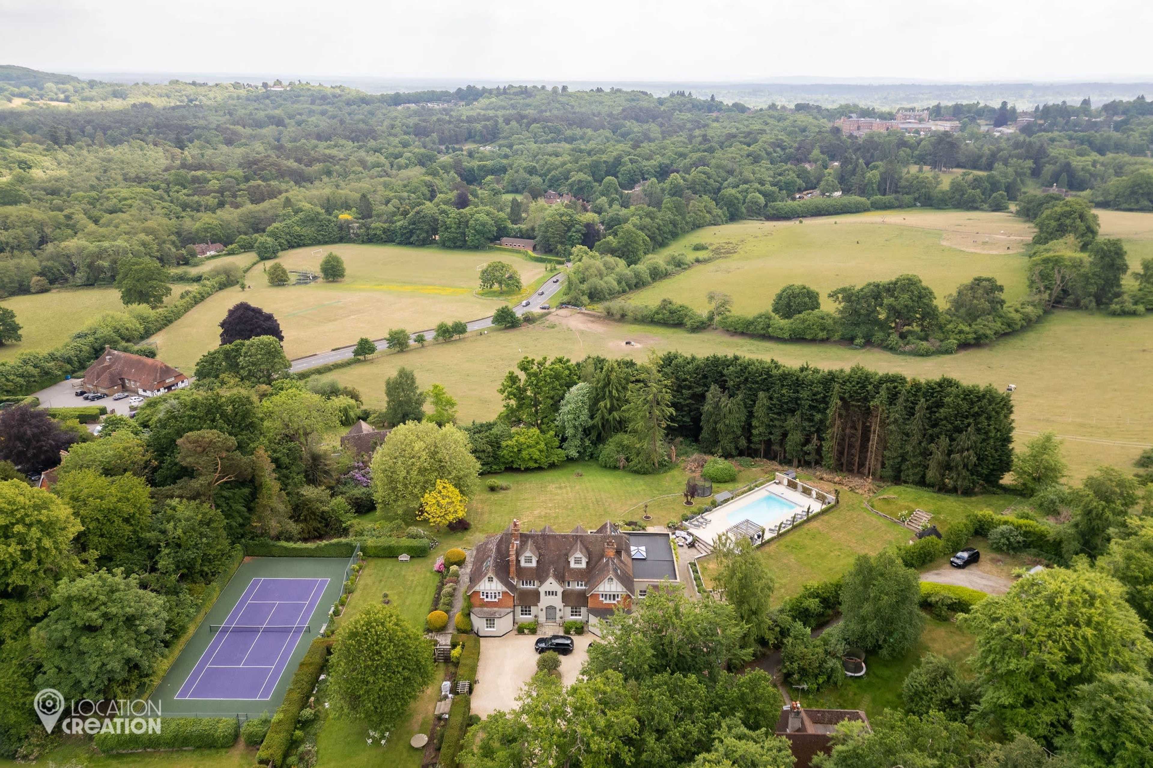 The image shows a large estate with a tennis court and swimming pool, surrounded by lush greenery and open fields.