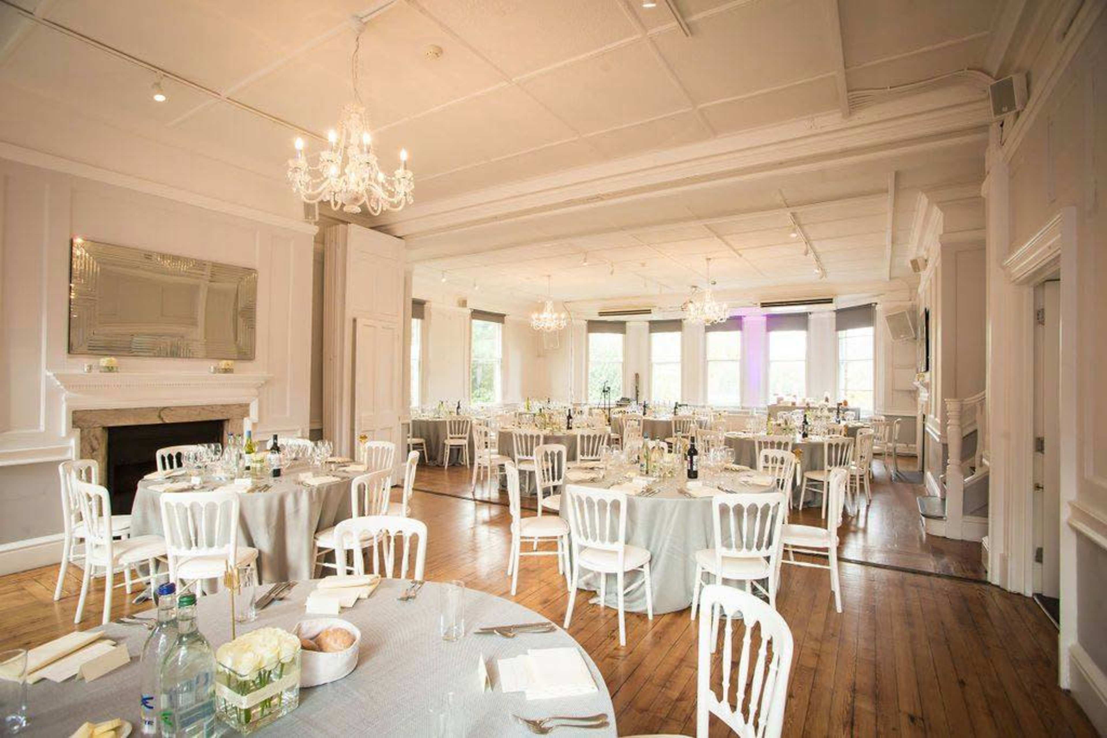 An elegantly arranged dining hall with round tables set for a formal event, featuring white chairs and chandeliers overhead.