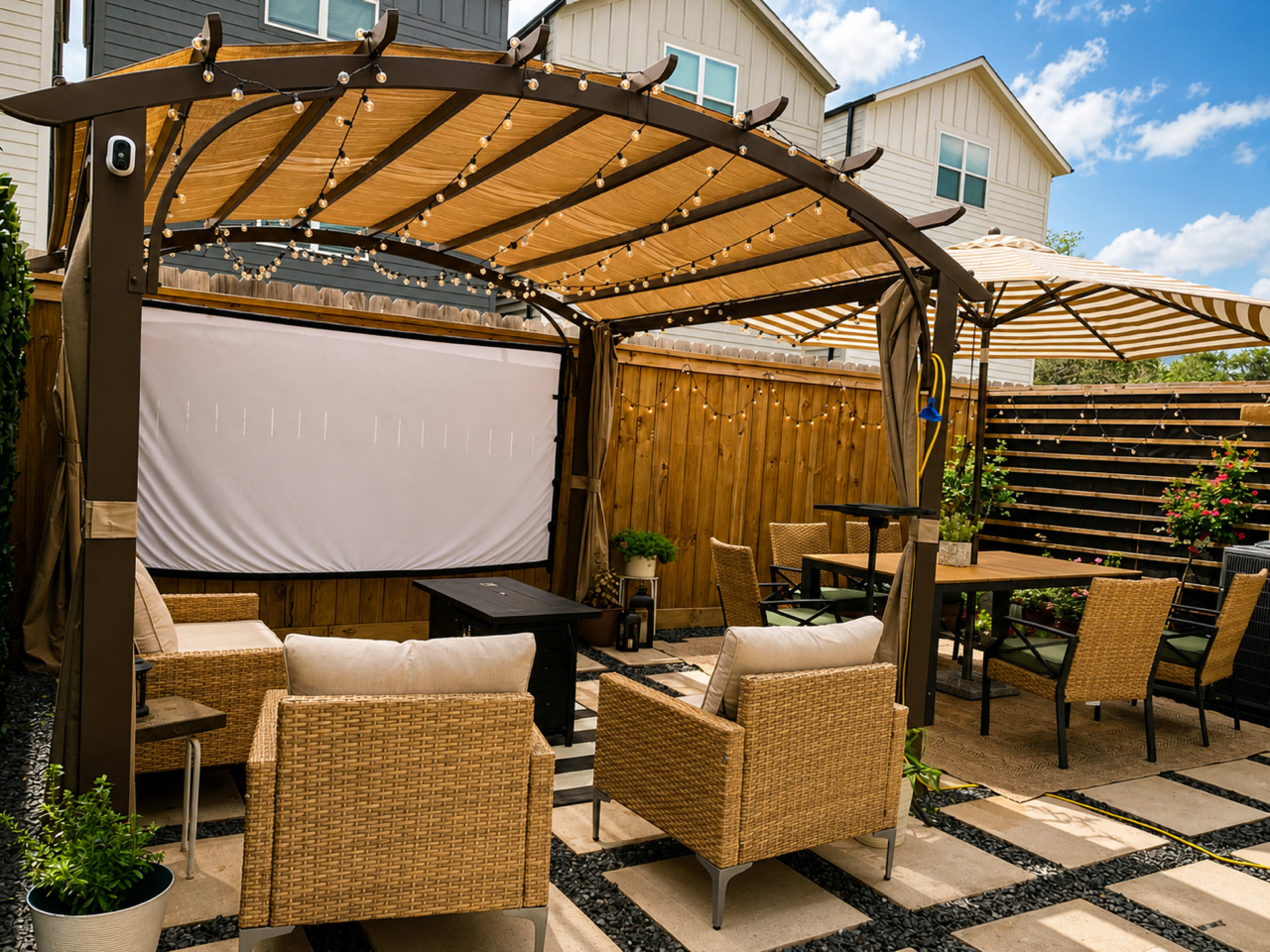 A cozy outdoor seating area features a pergola with string lights, a large screen, and various seating arrangements made of wicker furniture around a small coffee table.