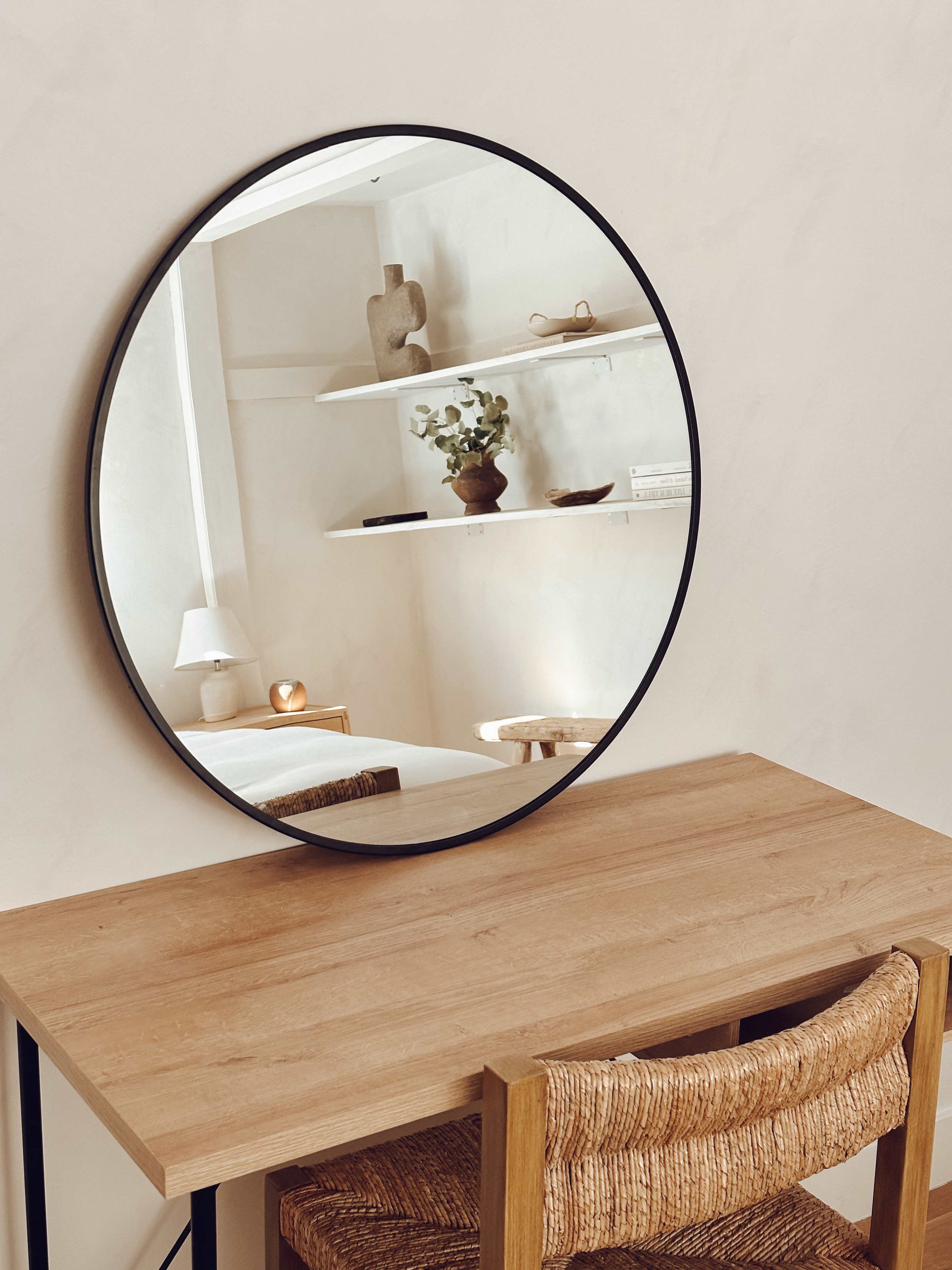 A round mirror reflects a wooden table and a shelf adorned with decor in a softly lit interior.