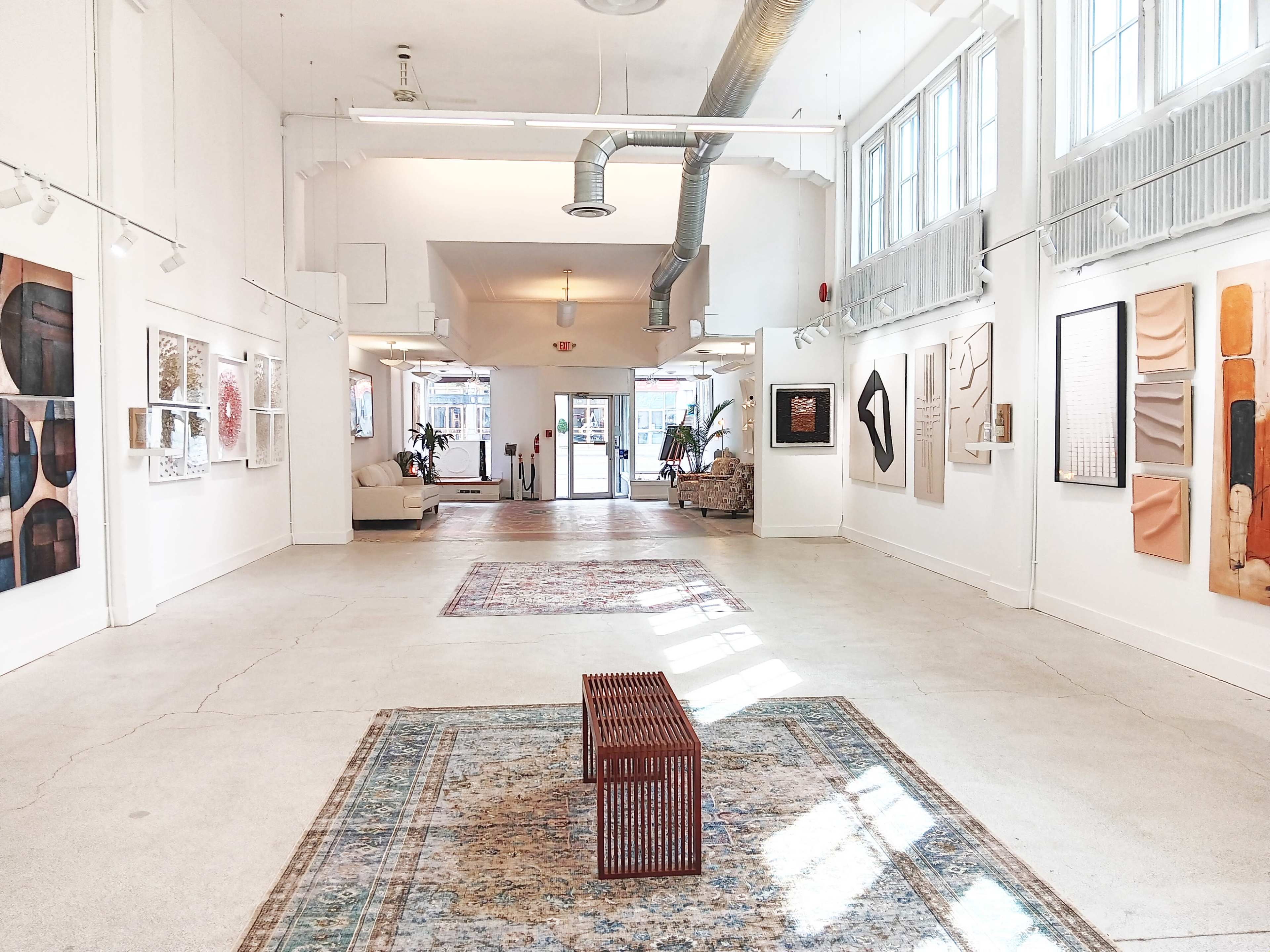 The image shows an art gallery interior with several framed artworks on the walls and a decorative wooden table in the center of the space.