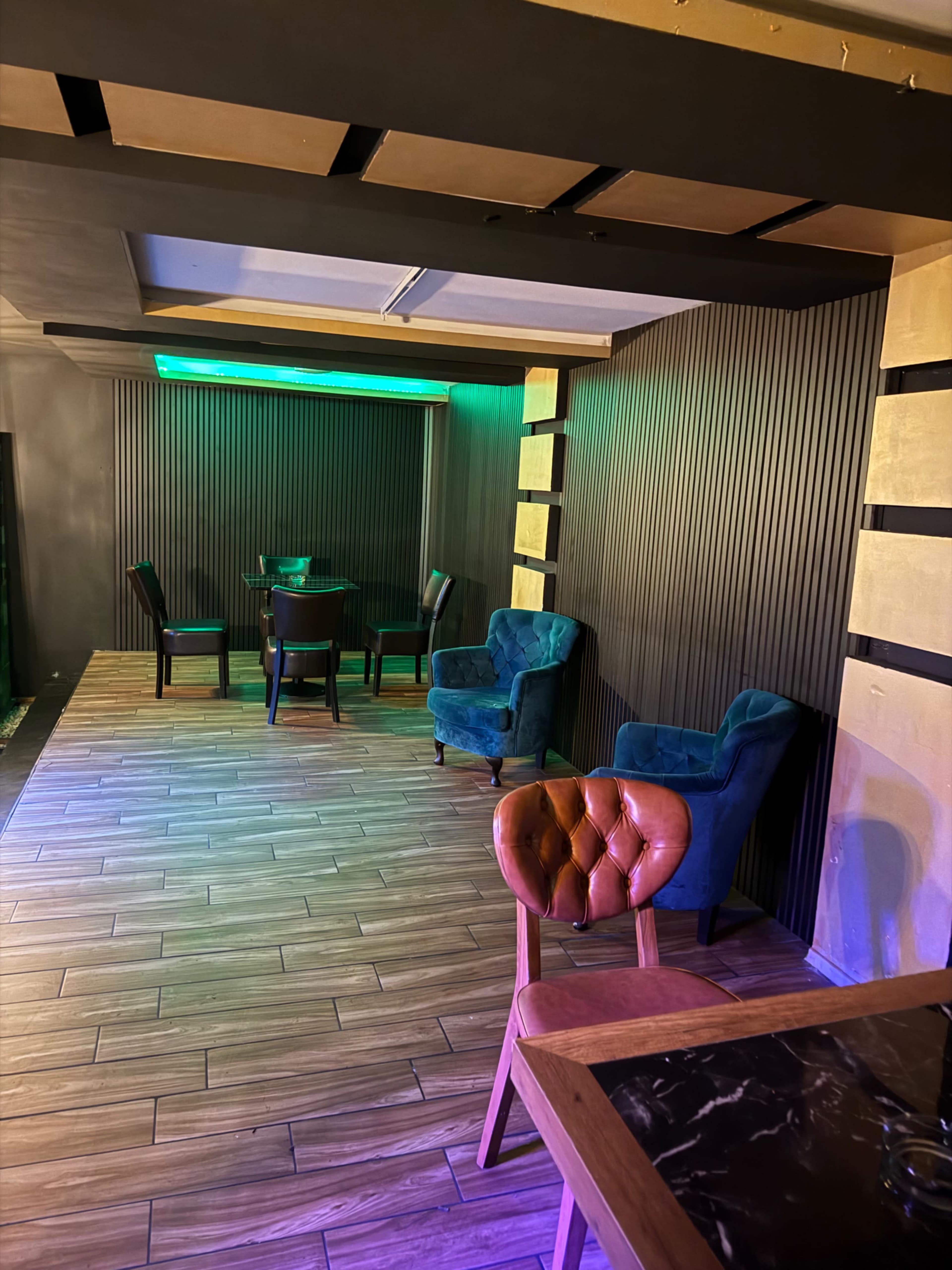 The image shows a modern interior space featuring a wooden floor, teal upholstered chairs, and a table arranged in a dimly lit area with colorful lighting.