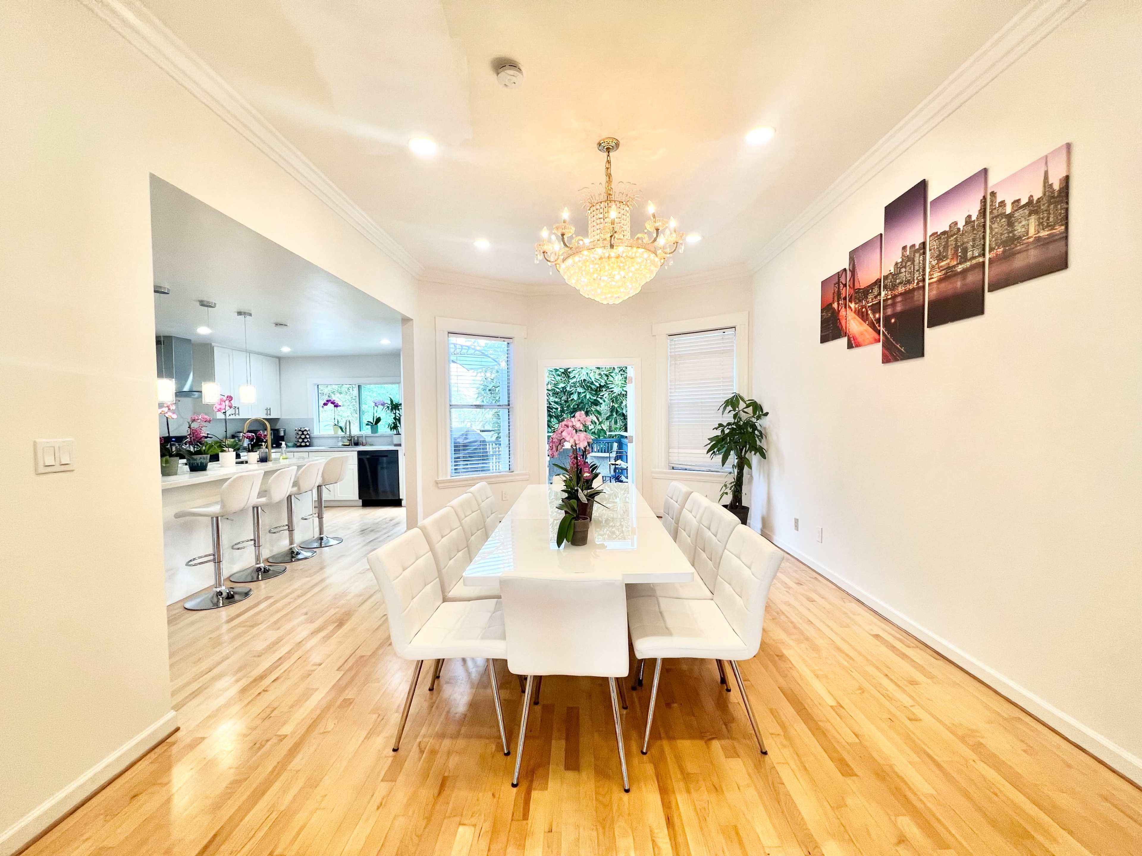 A modern dining room features a long white table surrounded by white chairs, with a chandelier above and a large window revealing kitchen views.