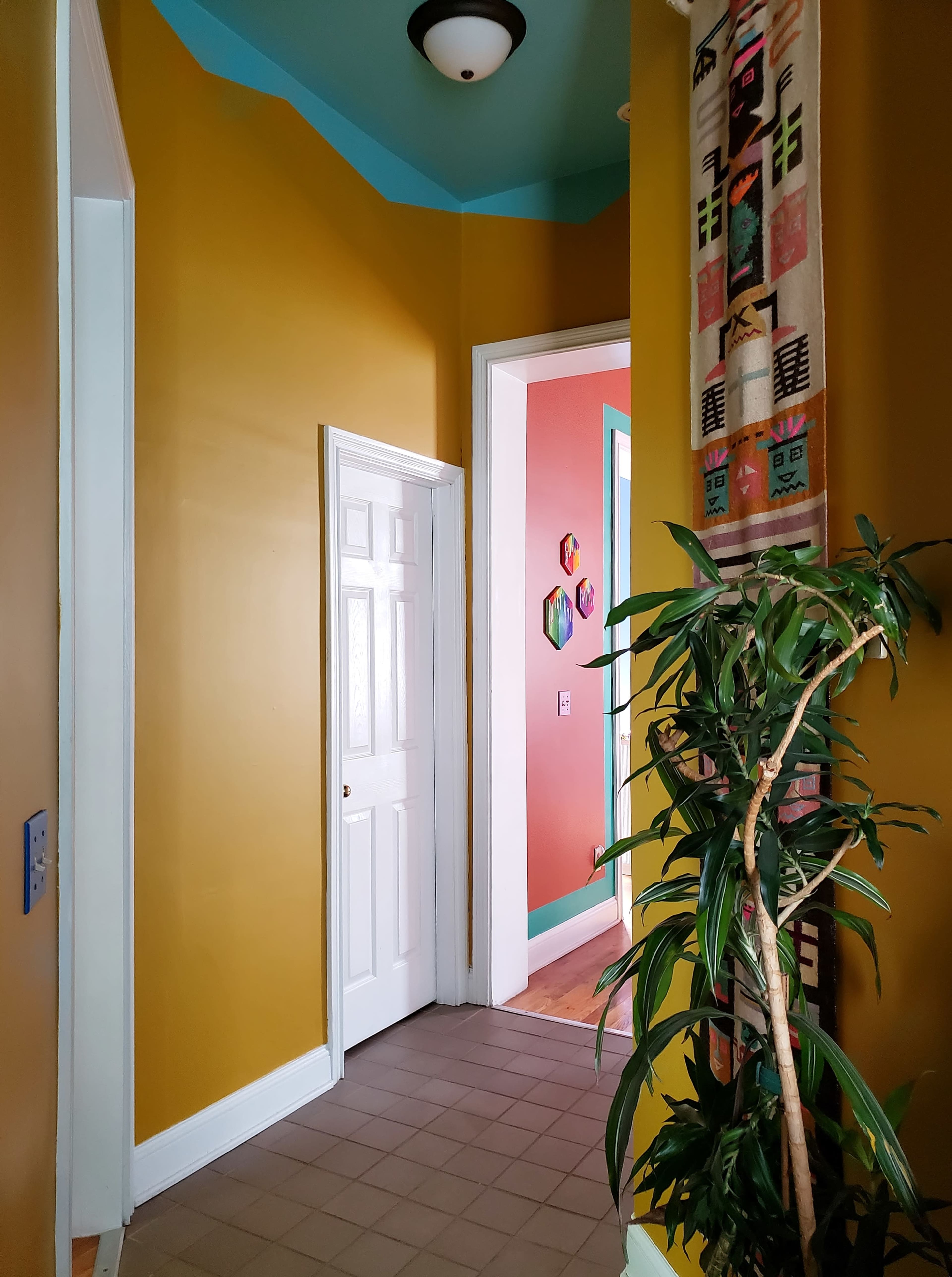 The image shows a hallway with yellow walls, a blue ceiling, and a doorway leading to a pink room, alongside a green plant in the foreground.