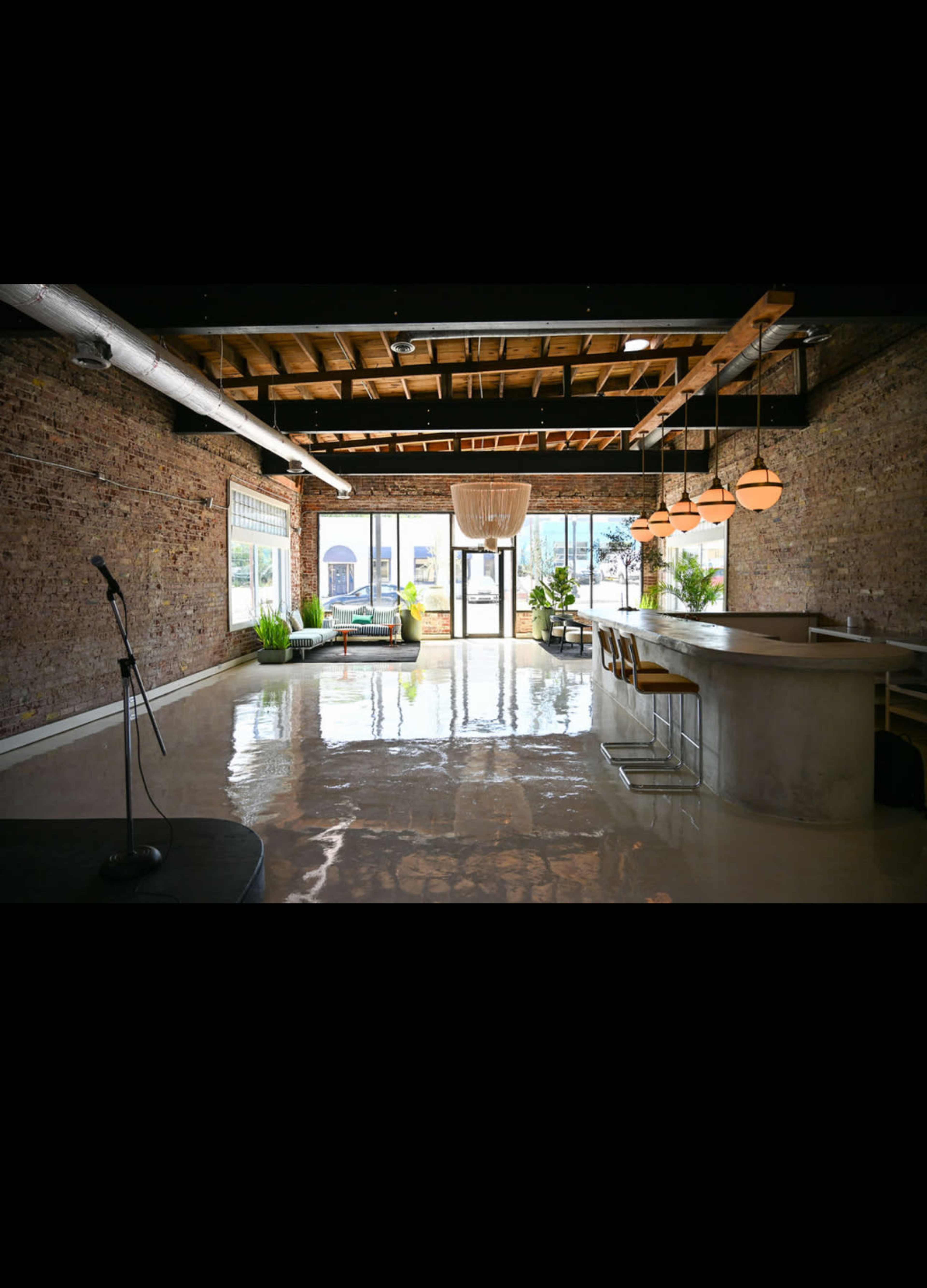 The image shows a spacious interior with exposed brick walls, a concrete bar, and polished floors, featuring a large chandelier and greenery.