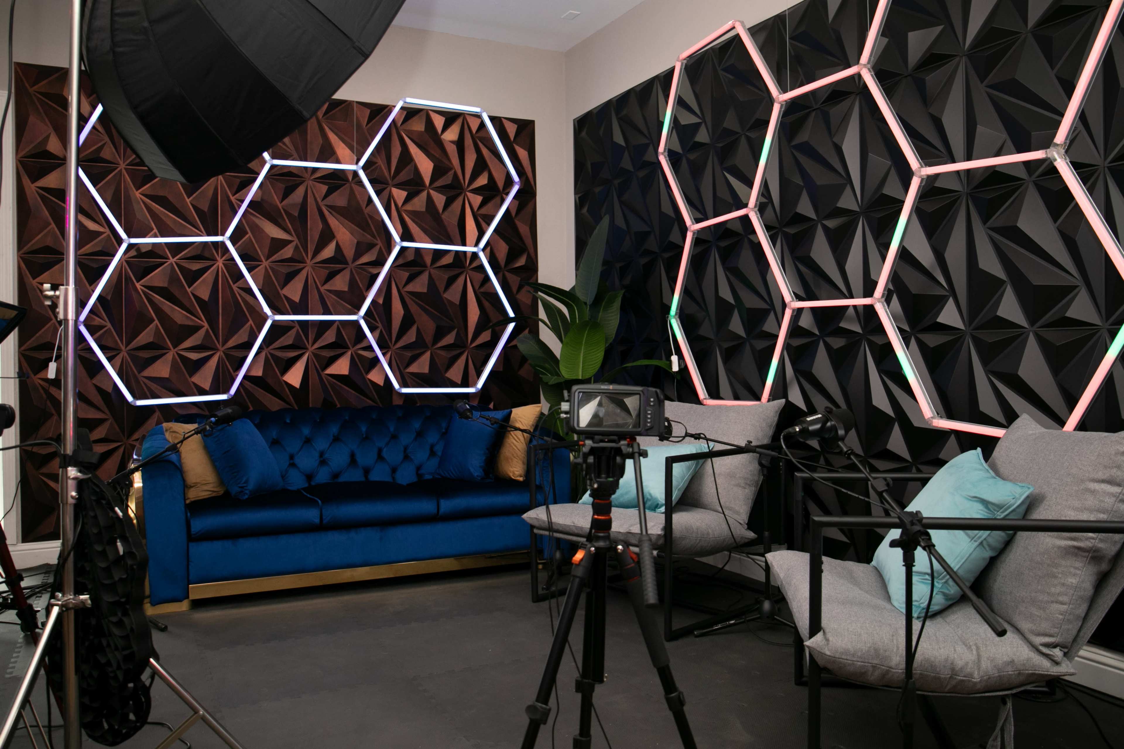 The image shows a modern interior studio setup with a blue sofa, geometric-patterned wall panels, and two gray chairs, accompanied by camera equipment and lighting.