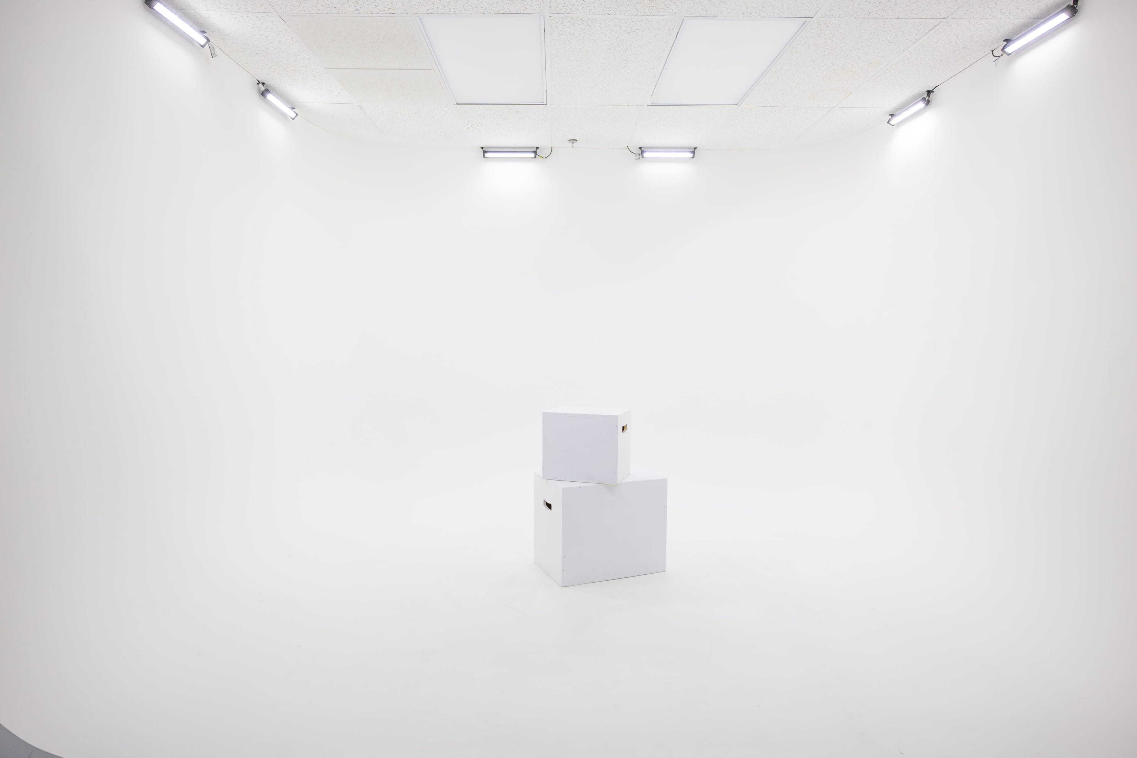 The image shows a sparsely furnished white studio space with two white boxes positioned on the floor.