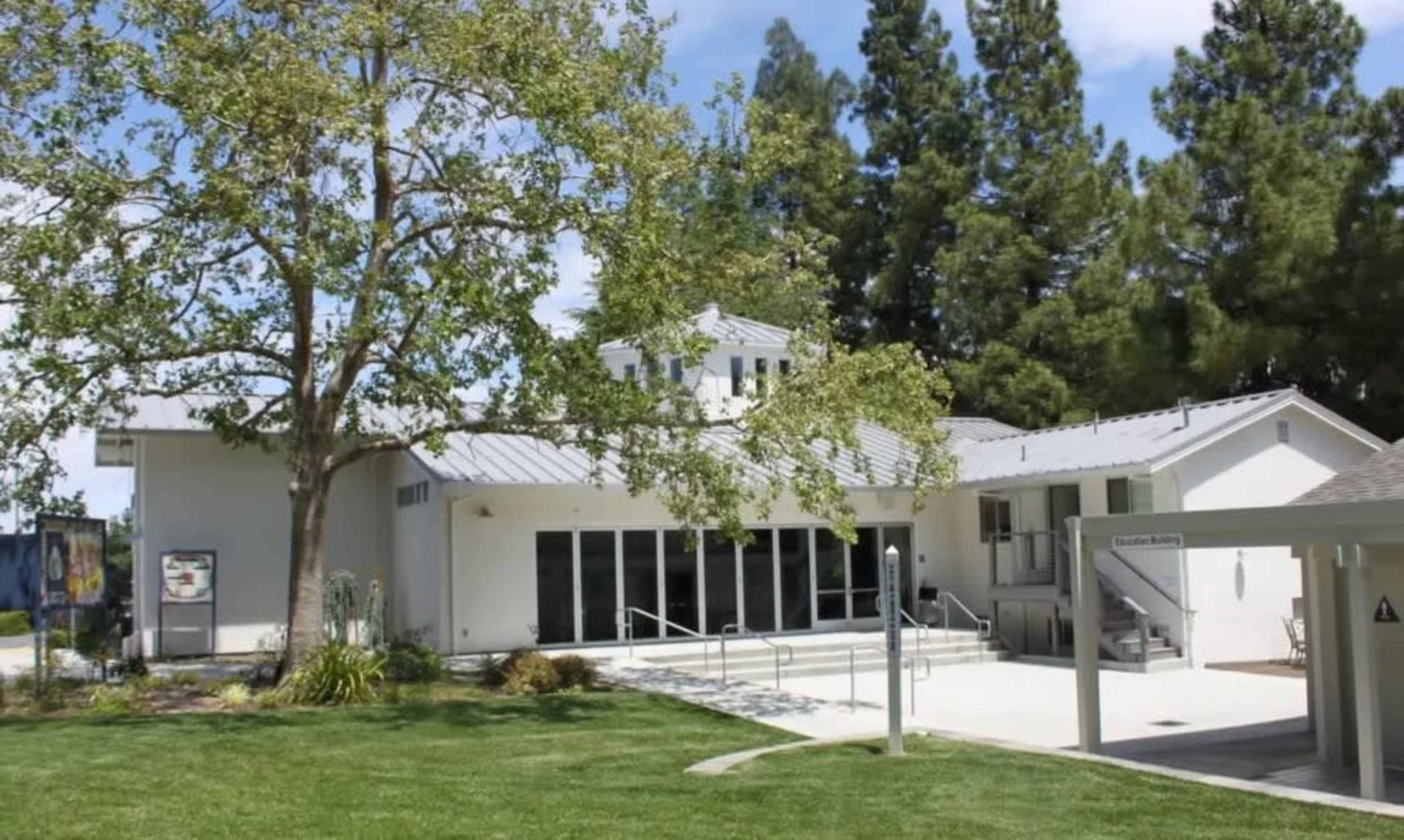 A modern building with large glass doors sits in a green landscape, surrounded by tall trees.