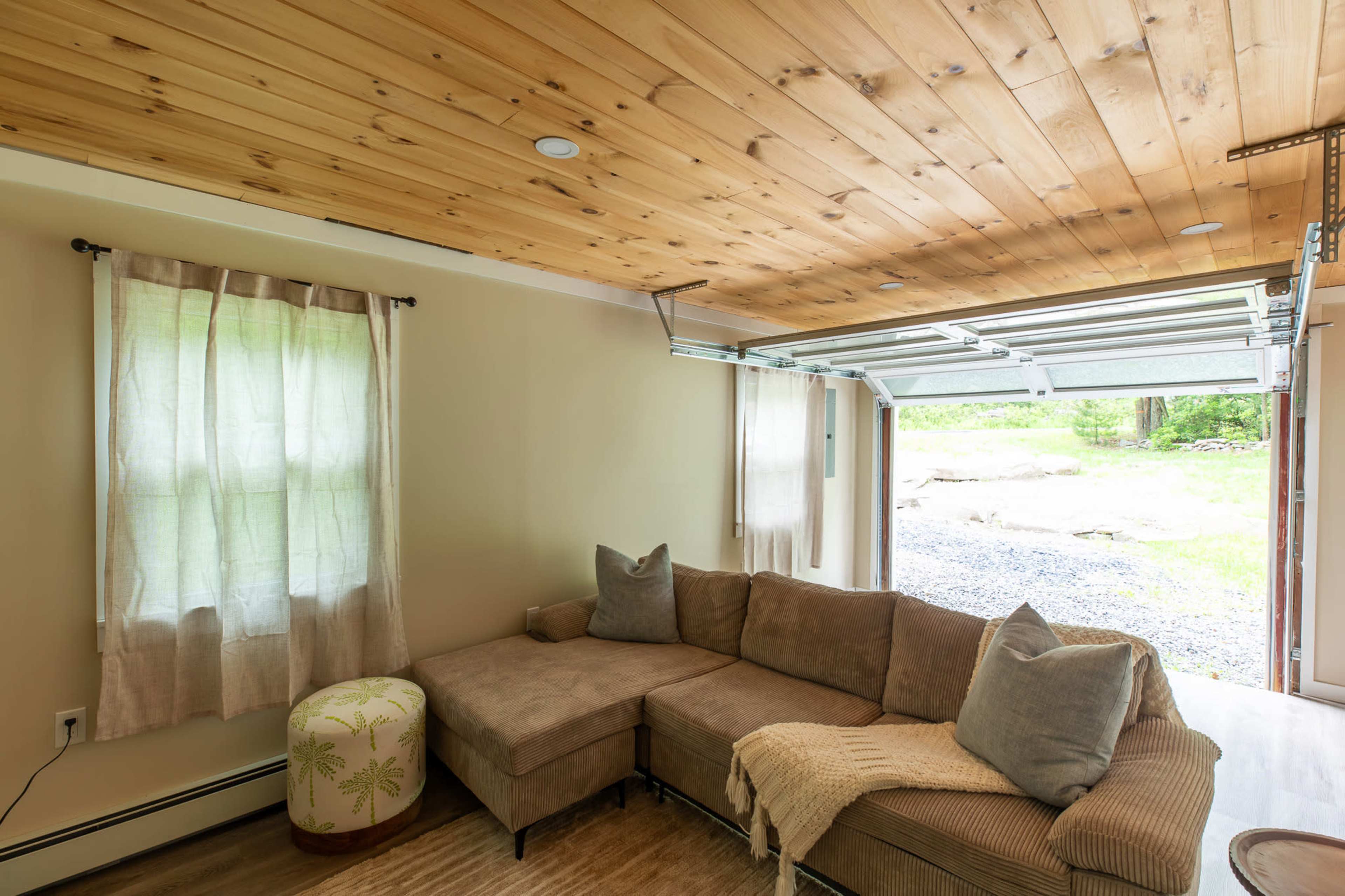 The image shows a living room with a beige sectional sofa, a light-colored knitted throw, a decorative stool, and a large garage door opening to a natural outdoor view.