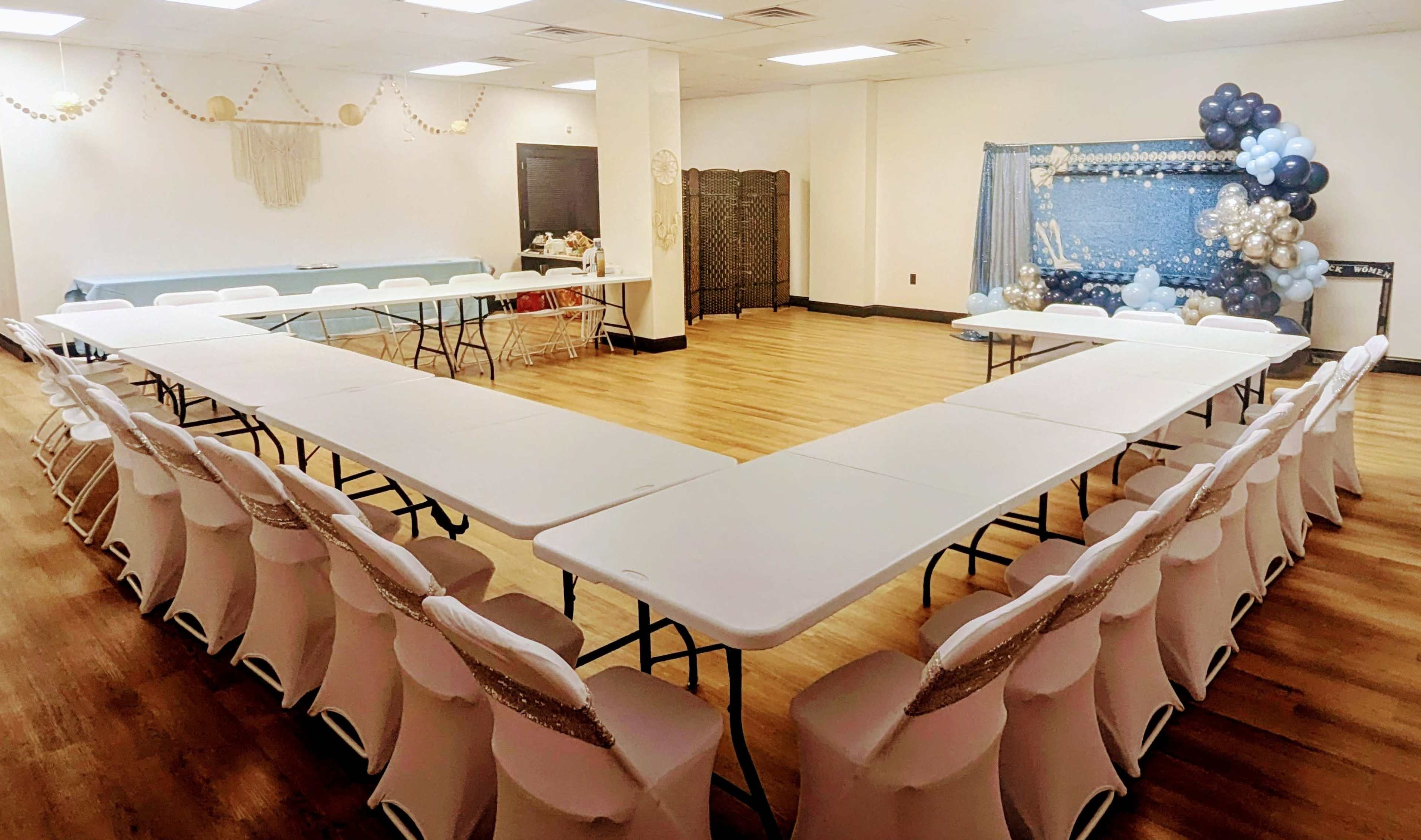 The image shows a decorated event space with multiple long tables arranged in a U-shape, surrounded by chairs, and a backdrop of balloons on one side.