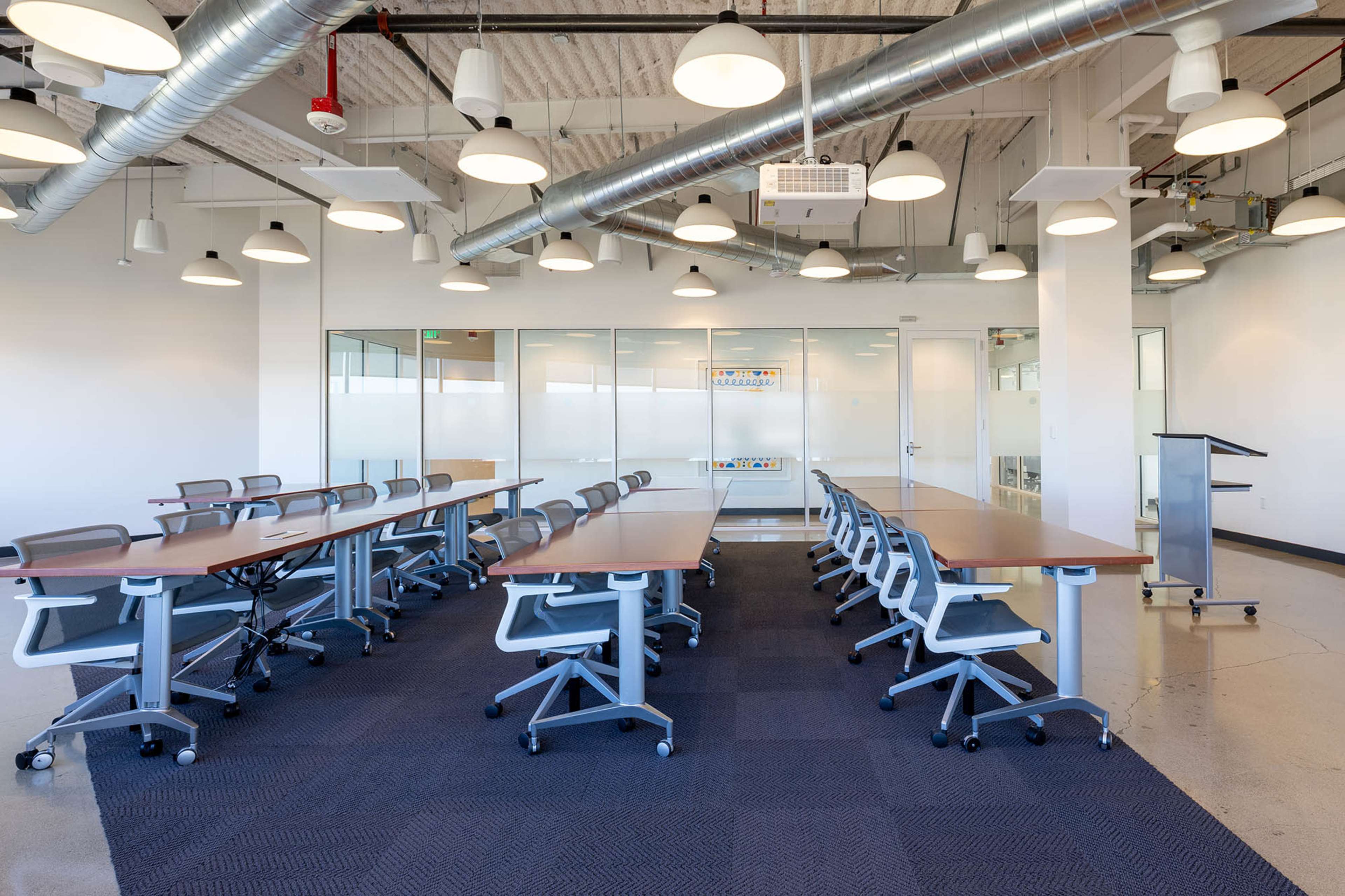 The image shows a modern conference room with several long tables arranged in a U-shape, surrounded by ergonomic chairs and illuminated by overhead lights.