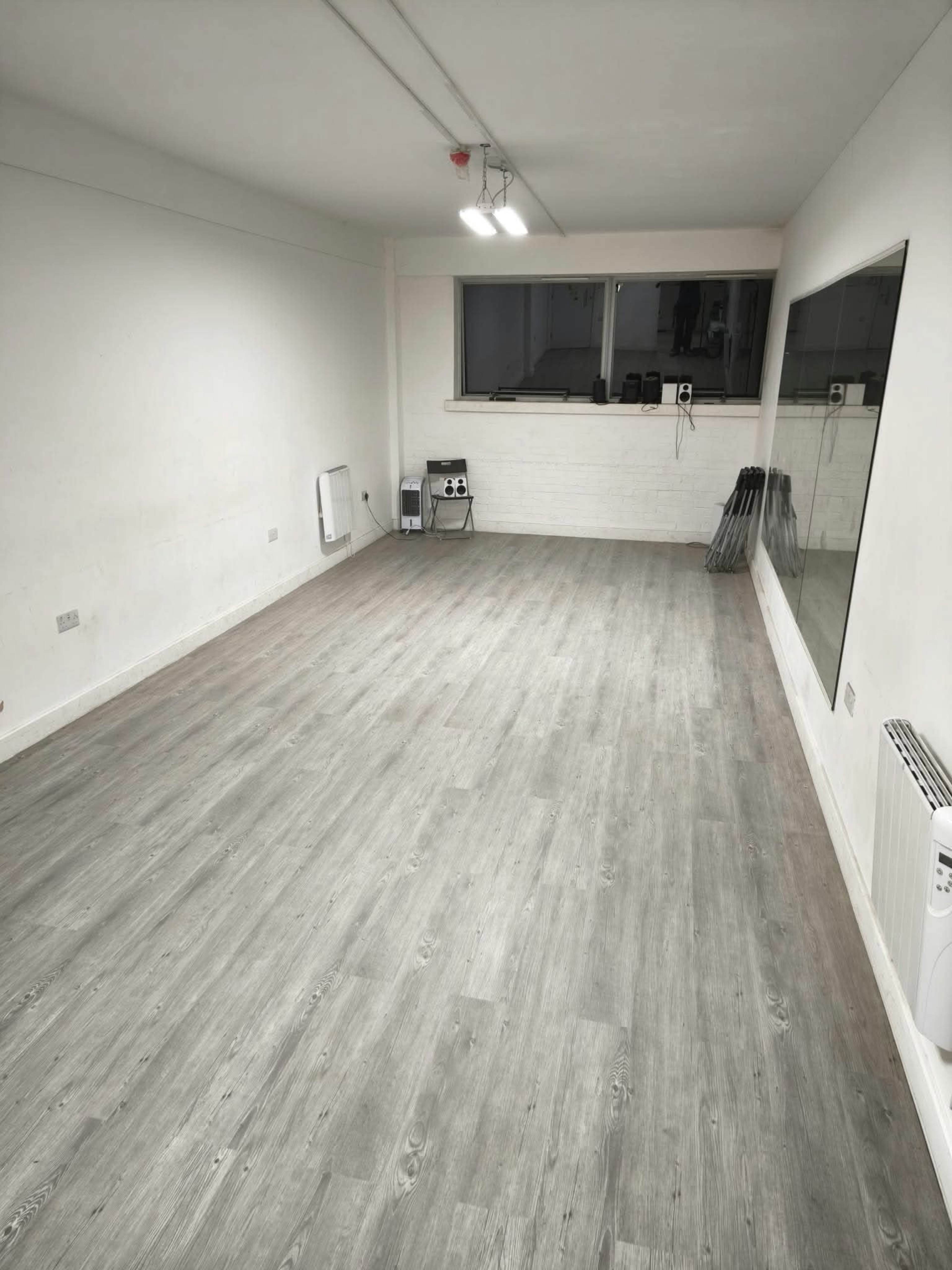 The image shows an empty room with wooden flooring, a large mirror on one wall, and a window above a small stack of equipment in the corner.