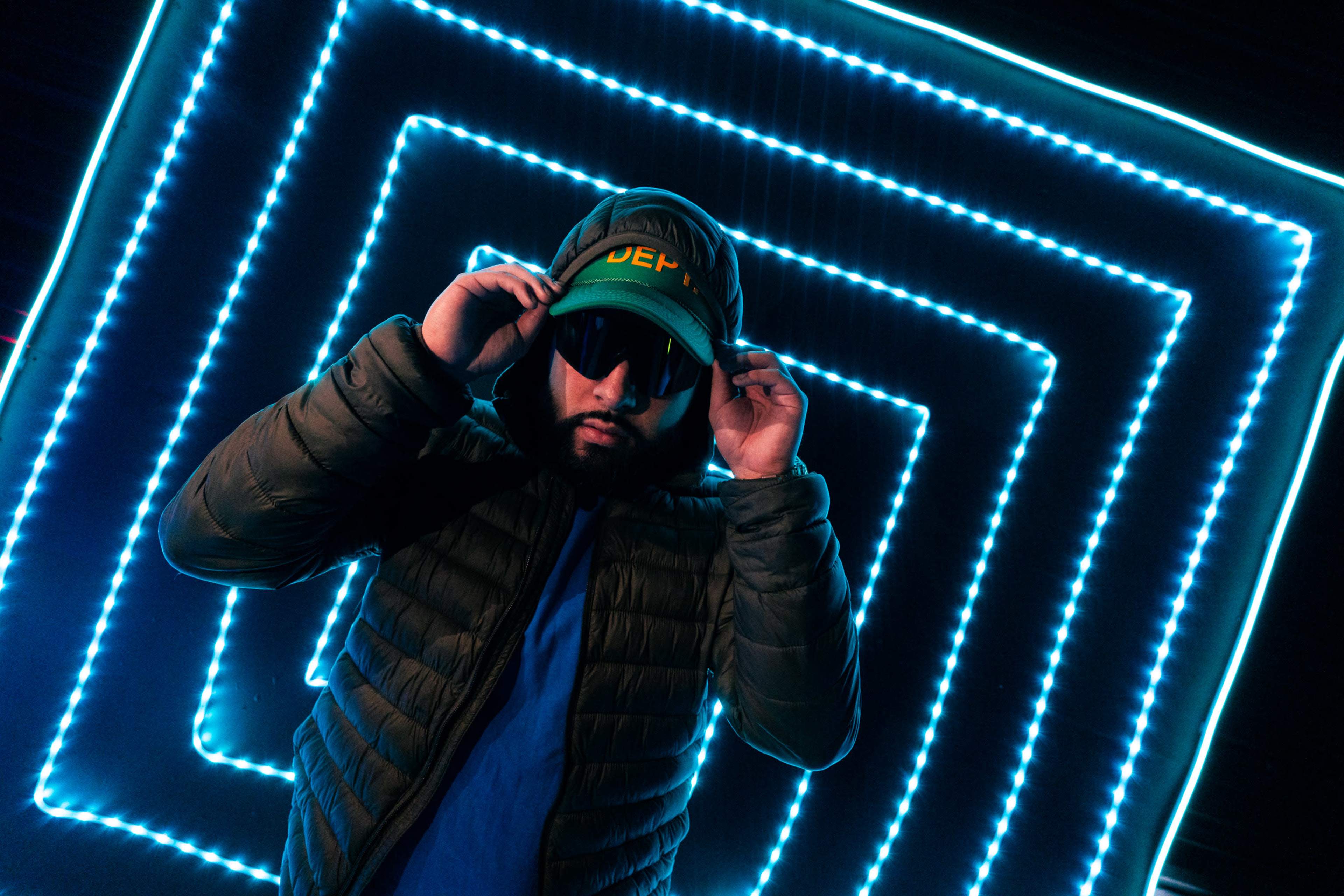A person wearing a green cap and sunglasses poses in front of a bright, glowing, geometric pattern.
