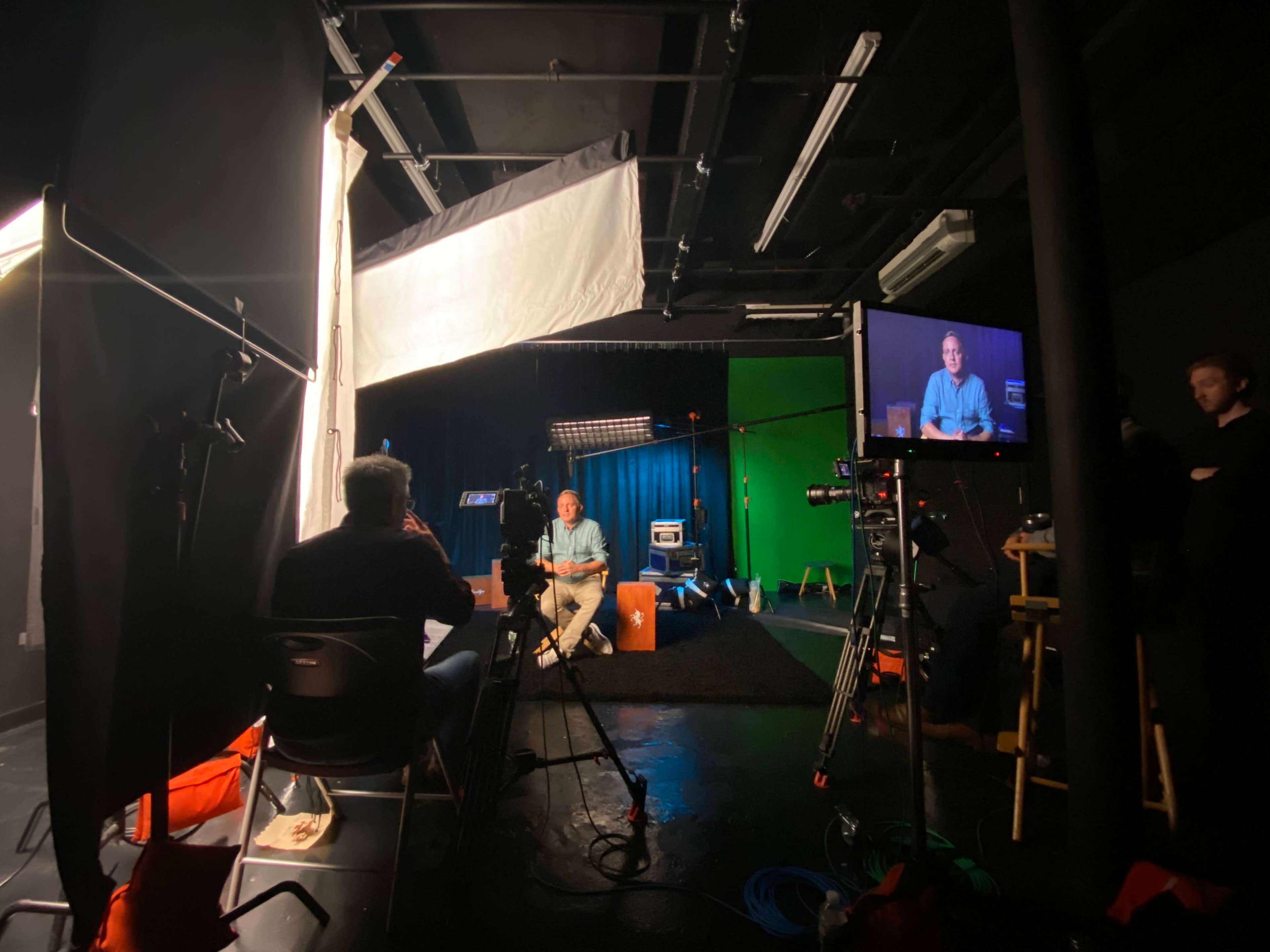 A studio setup features a speaker sitting on a stool with cameras and lighting equipment arranged around them.