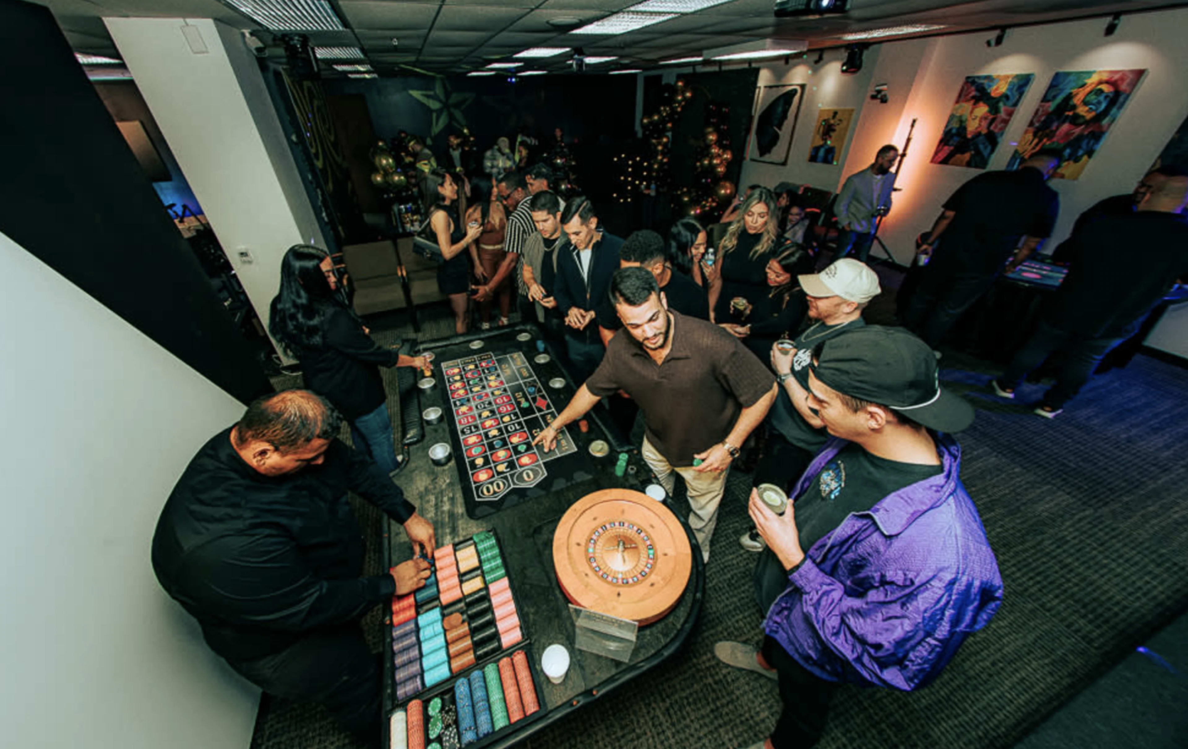 A group of people gathers around a roulette table in a festive indoor setting, with decorations in the background.