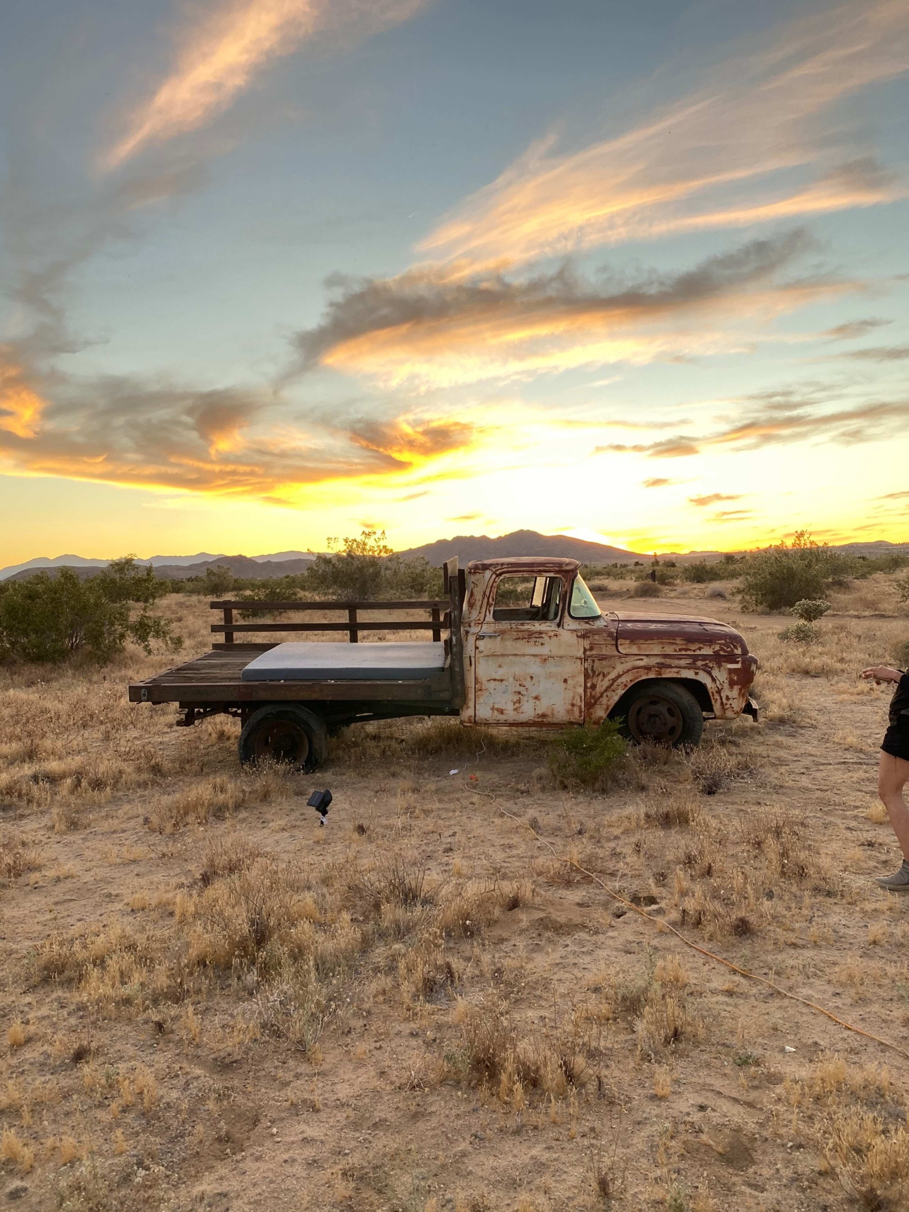 A rusty vintage truck is parked on a desert landscape with a vibrant sunset in the background.