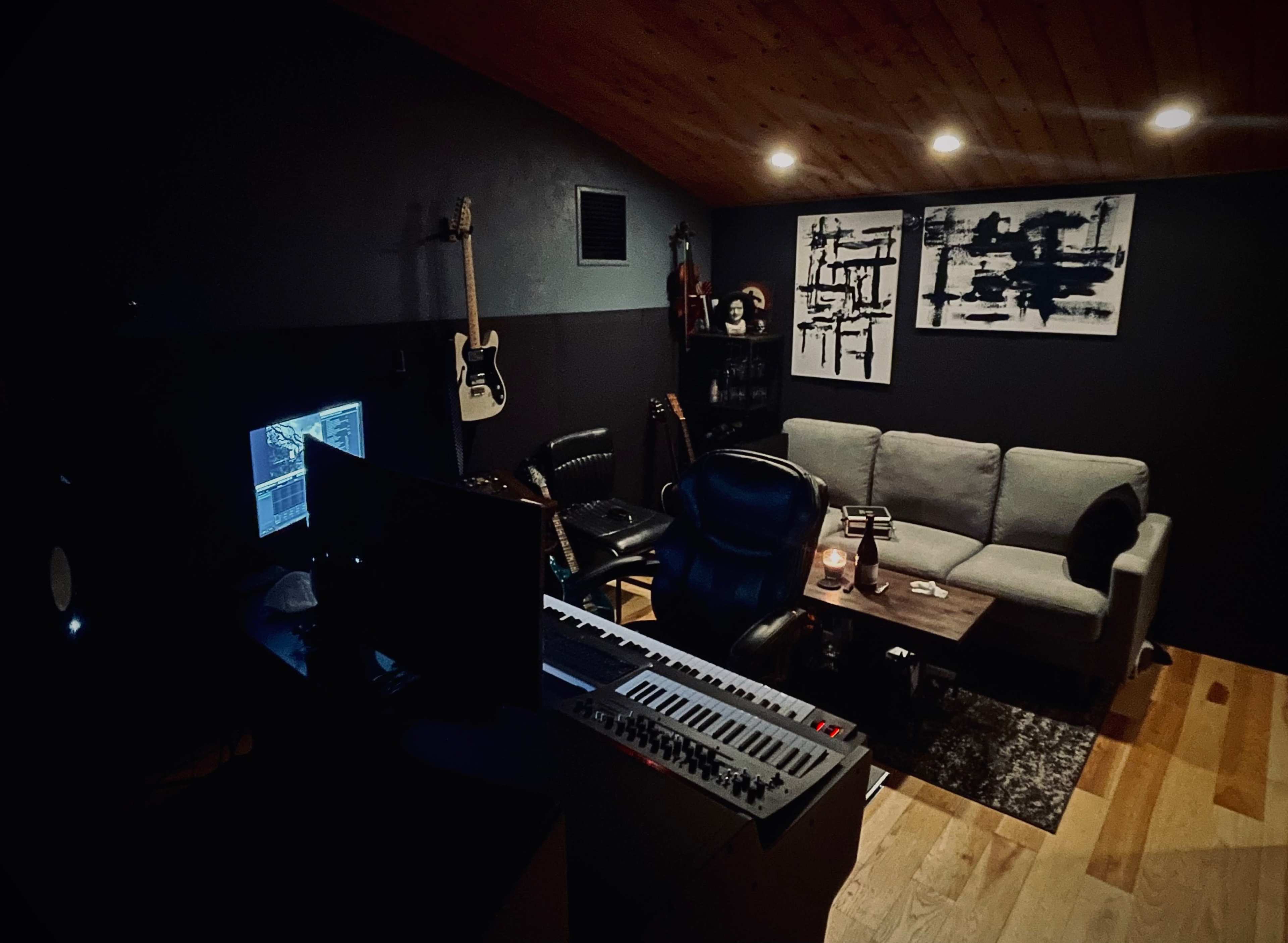 A dimly lit music studio featuring a keyboard, a desk with a computer monitor, a gray sofa, a wooden table, and guitars mounted on the wall.