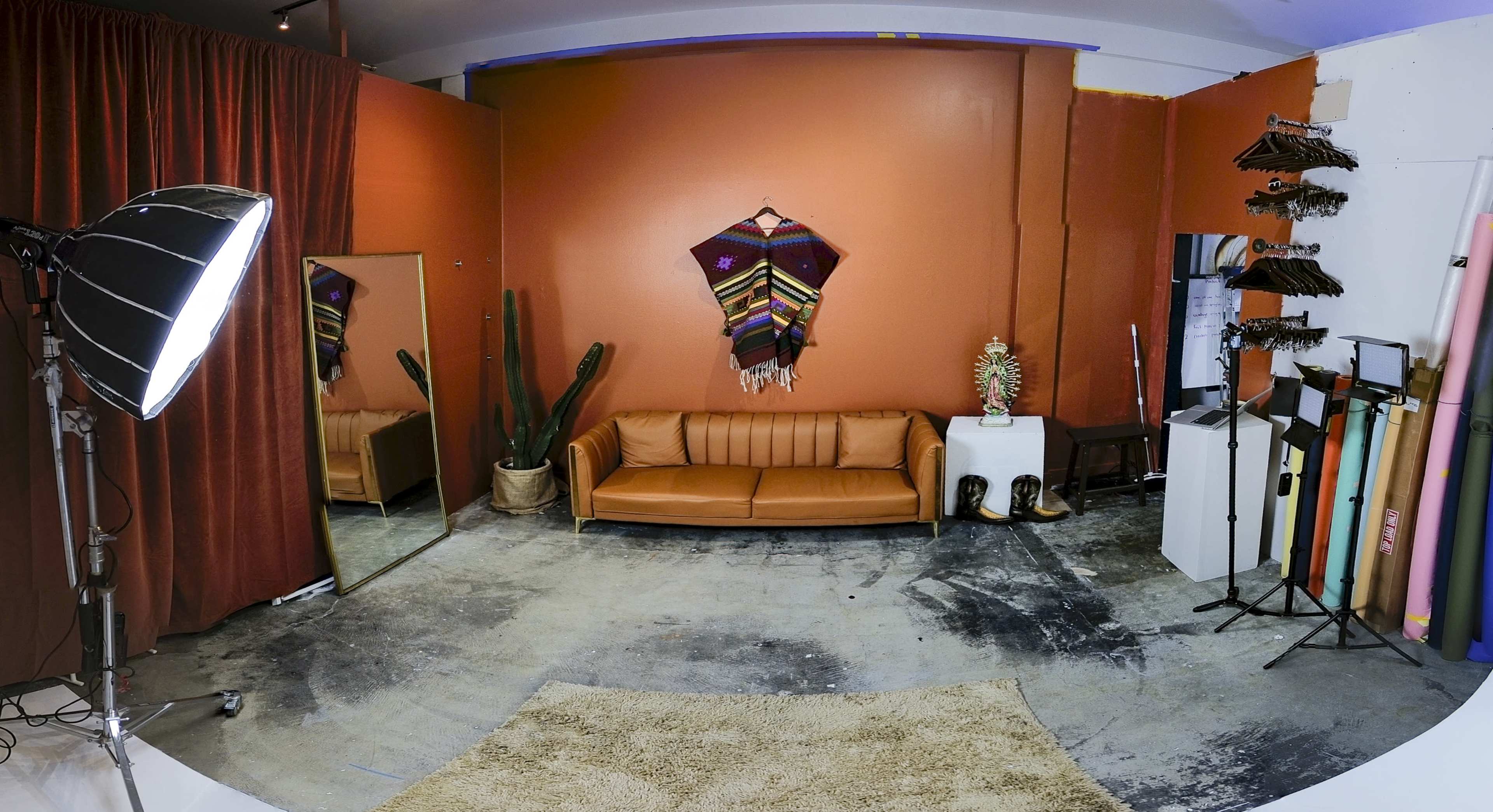 The image shows a studio space featuring an orange wall, a brown leather sofa, a large mirror, a potted cactus, and racks with clothing hangers, alongside photography equipment and colorful backdrops.