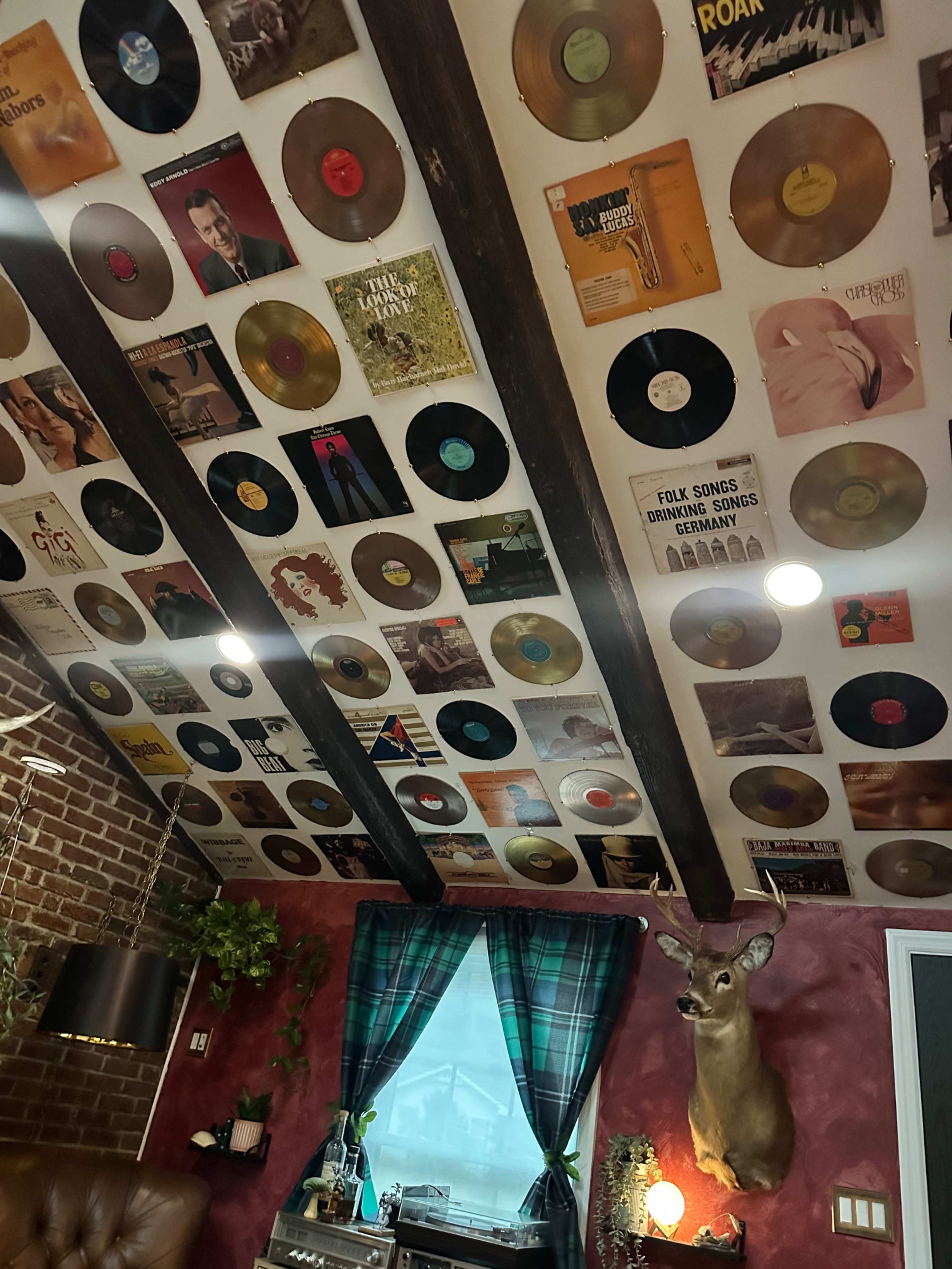 The ceiling is adorned with various vinyl records and album covers, while a mounted deer head decorates the wall beneath a window with green checked curtains.