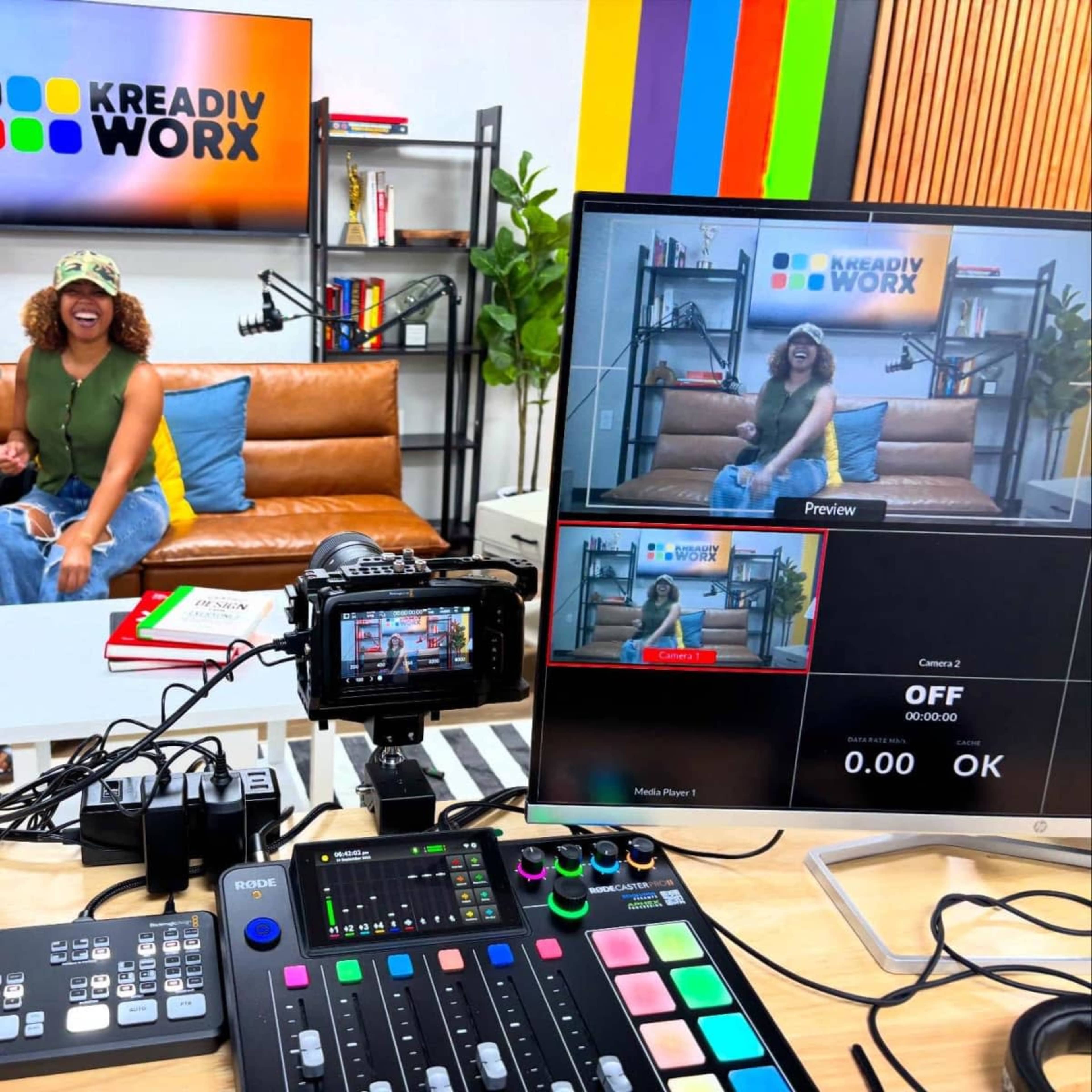 A television studio setup features a host in casual attire sitting on a couch, with a control panel and monitors displaying a live video feed.