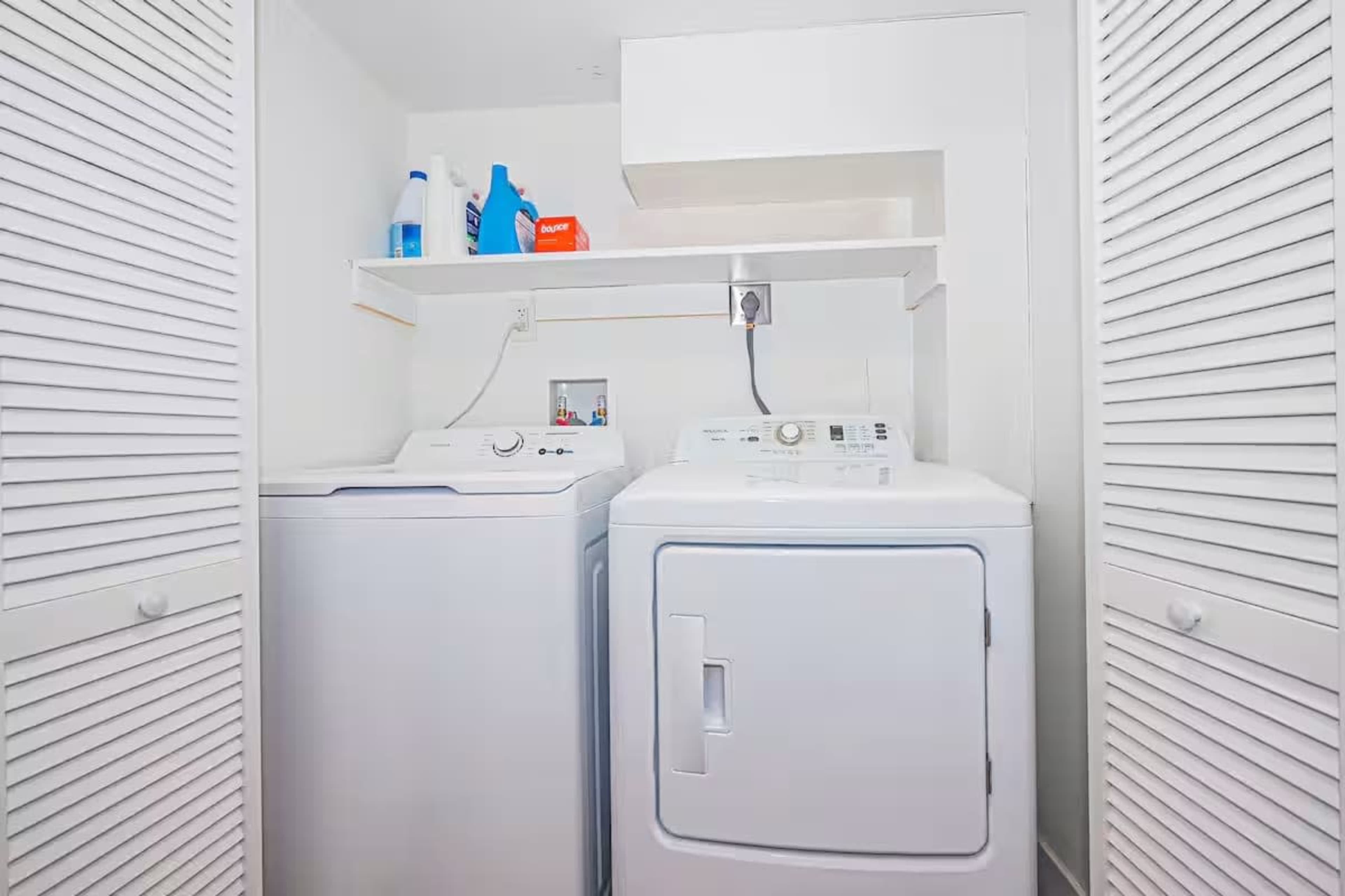 A laundry room is shown with a washing machine and dryer side by side, along with shelves and cleaning supplies on a wall above.
