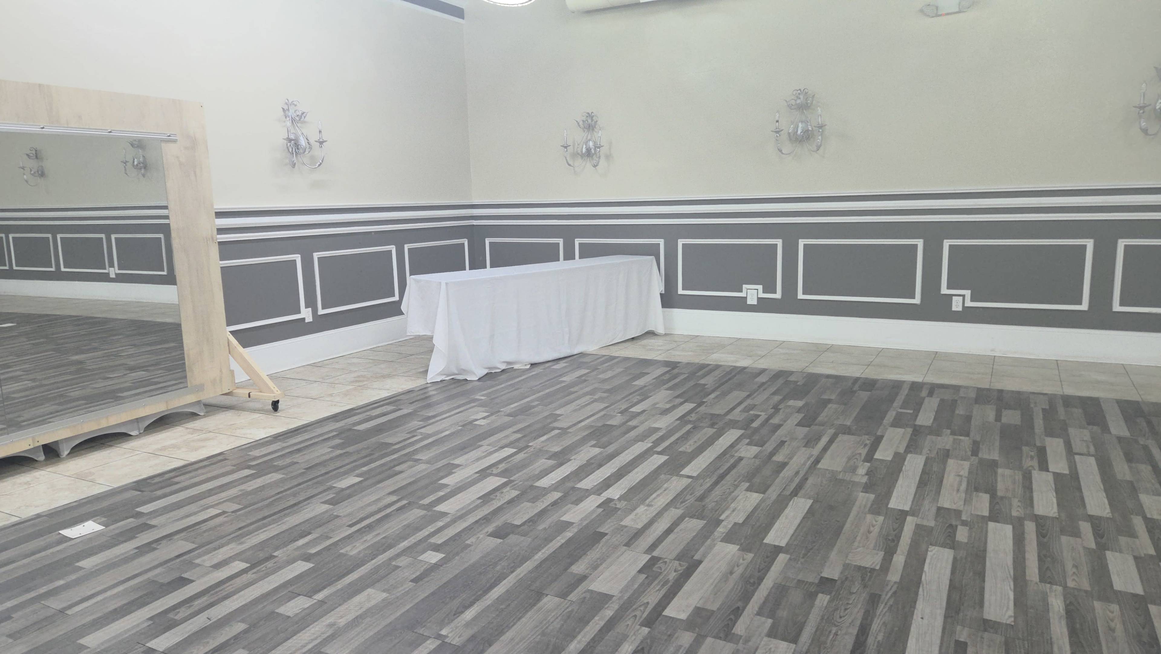 The image shows a spacious room with a tiled floor, gray walls adorned with decorative molding, a table covered with a white cloth, and a large mirror on an easel.