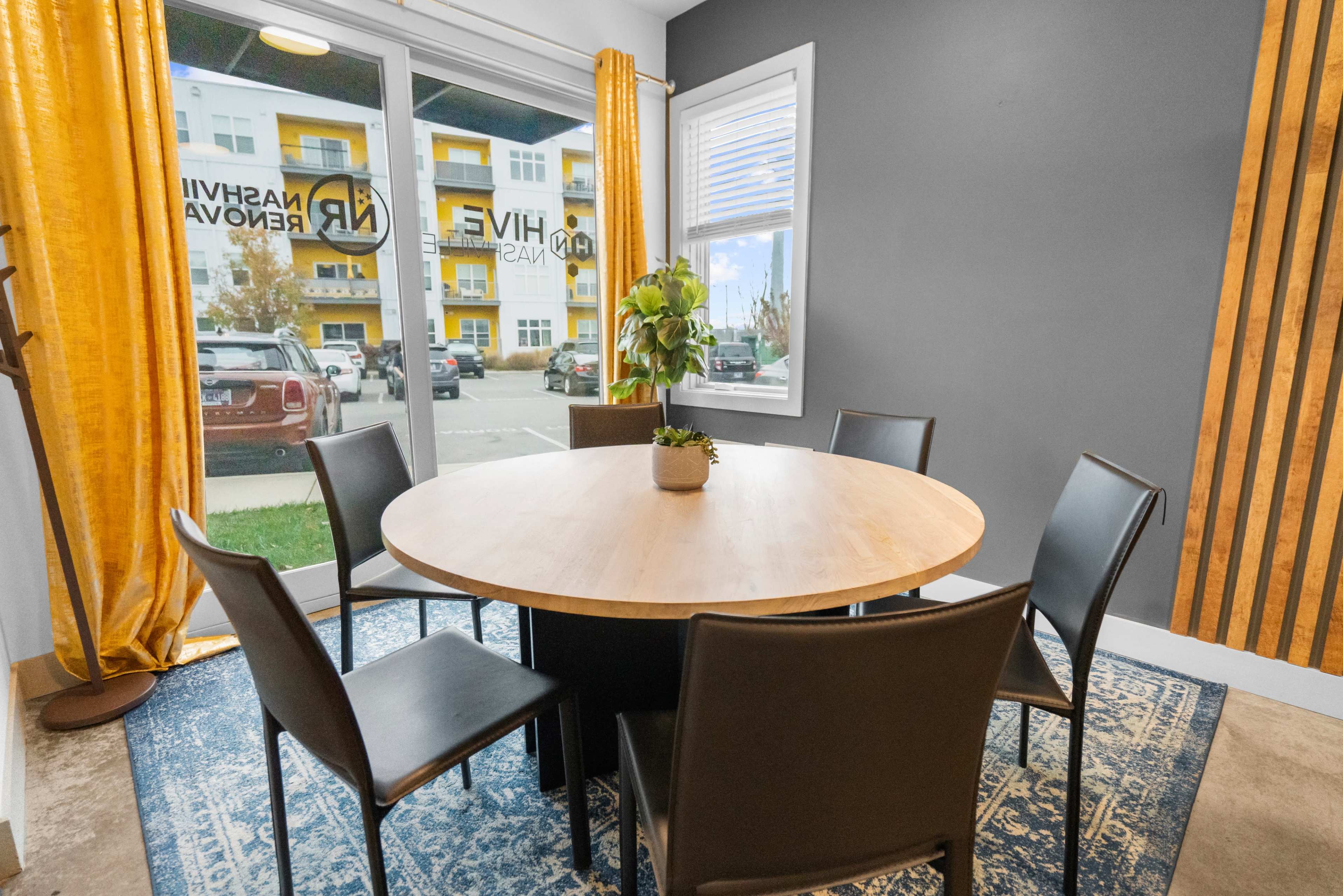 A round table with six black chairs is set in a modern room with large windows and yellow curtains.