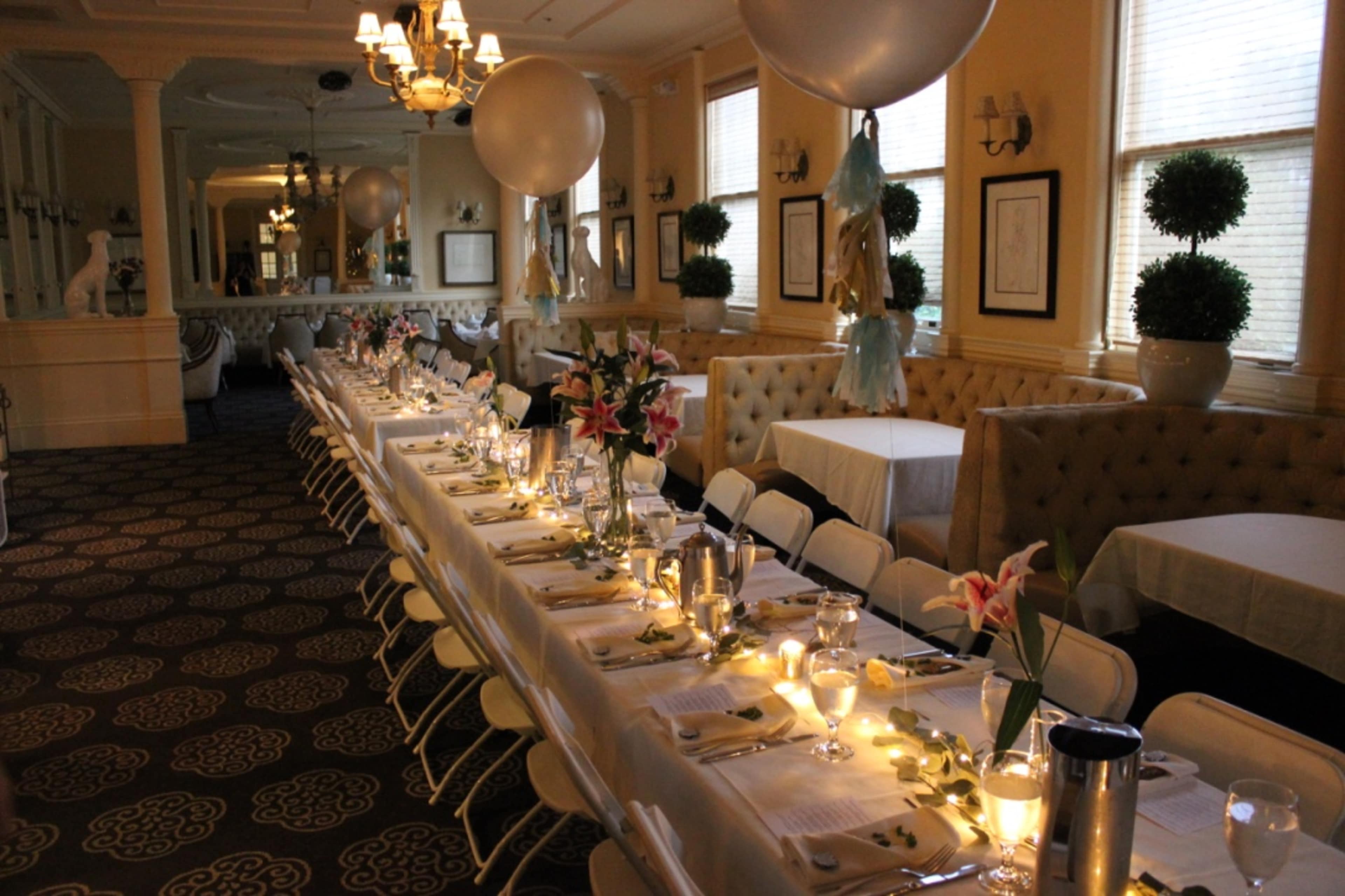 A long, elegantly set dining table with floral arrangements and candles is displayed in a well-decorated room featuring soft lighting and balloon decorations.