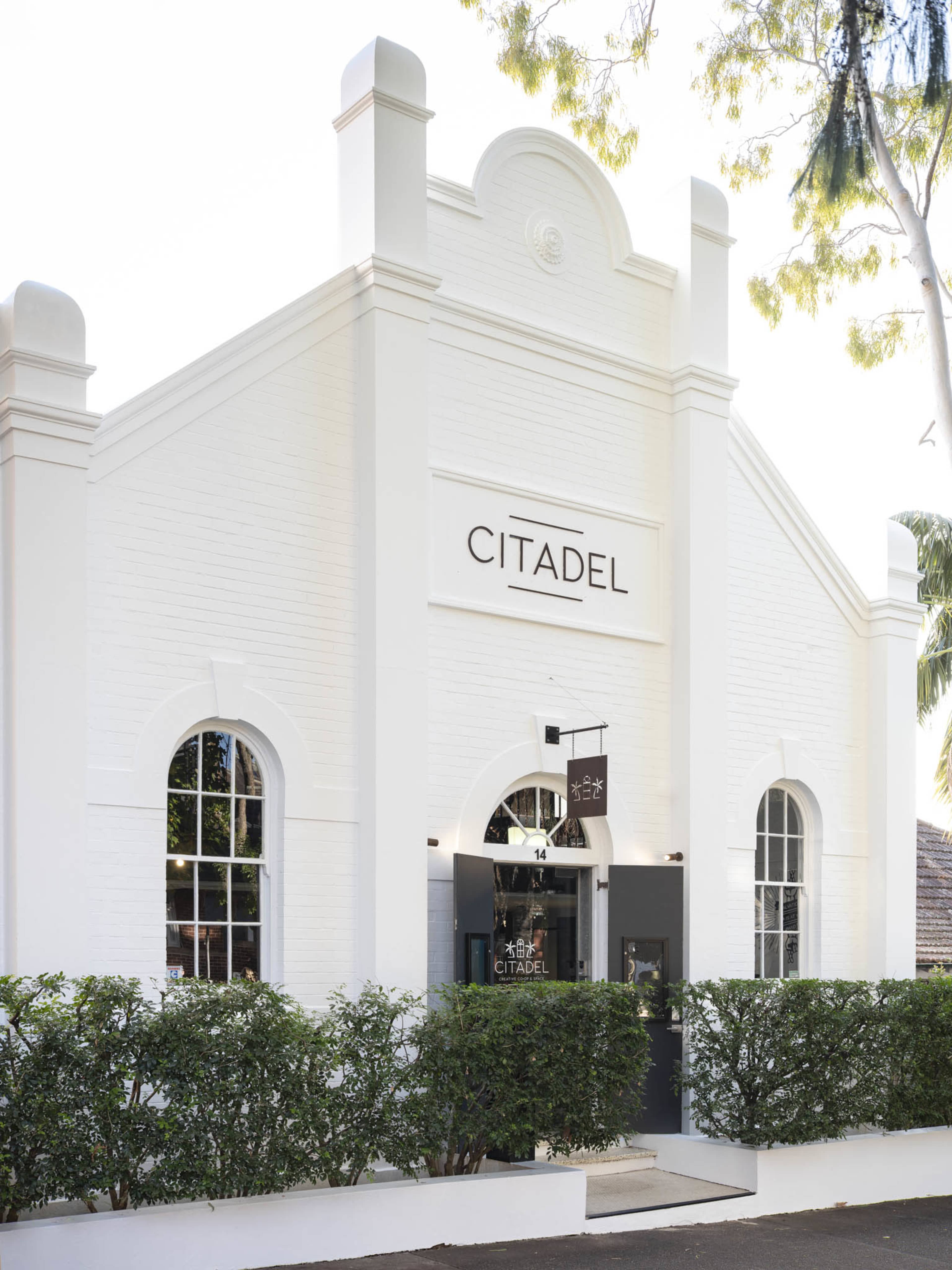 The image shows a white, modern building with large windows and a sign reading "CITADEL," surrounded by neatly trimmed hedges.