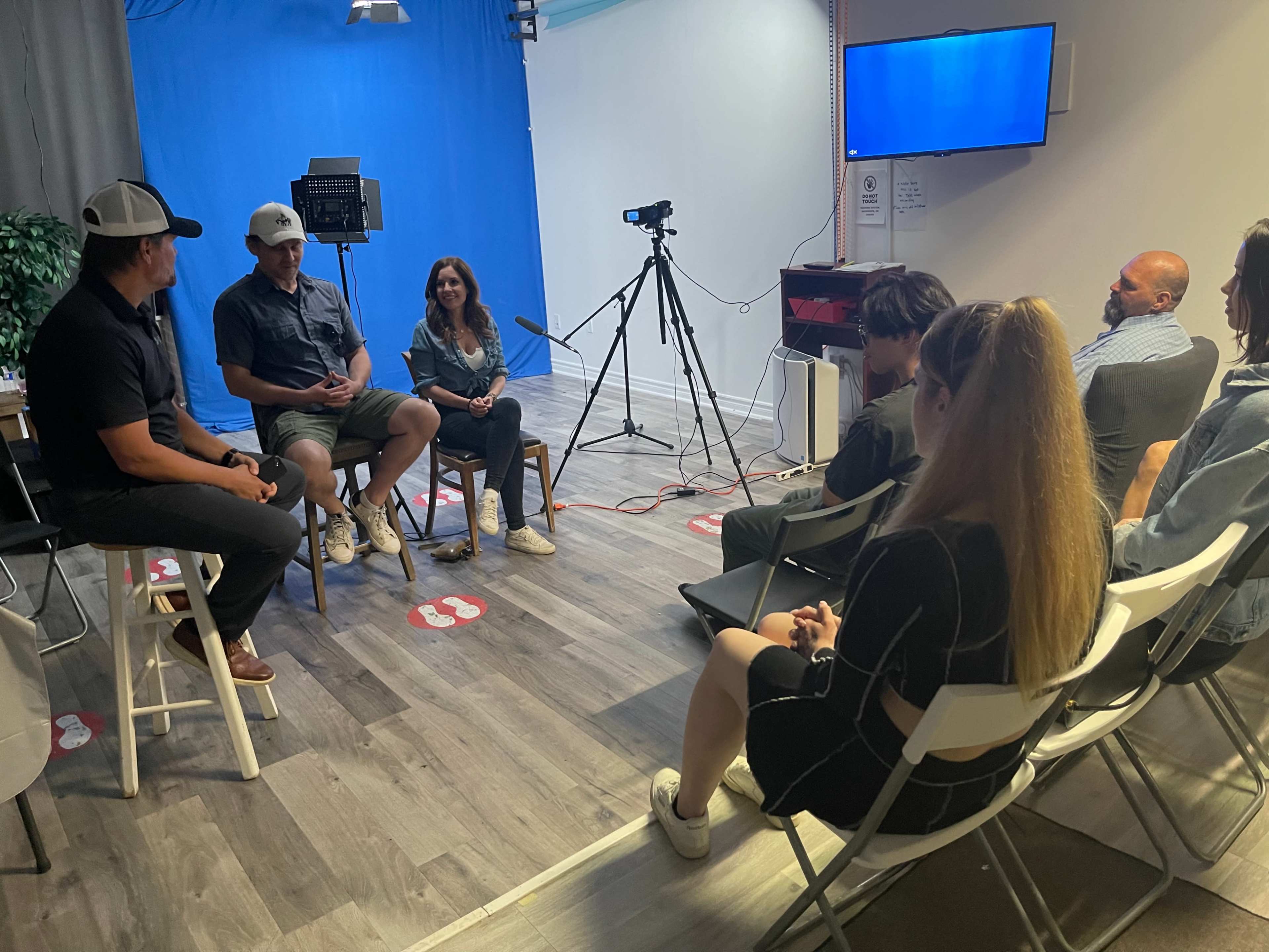 A group of people sits in chairs facing a small stage with a blue backdrop and camera equipment.