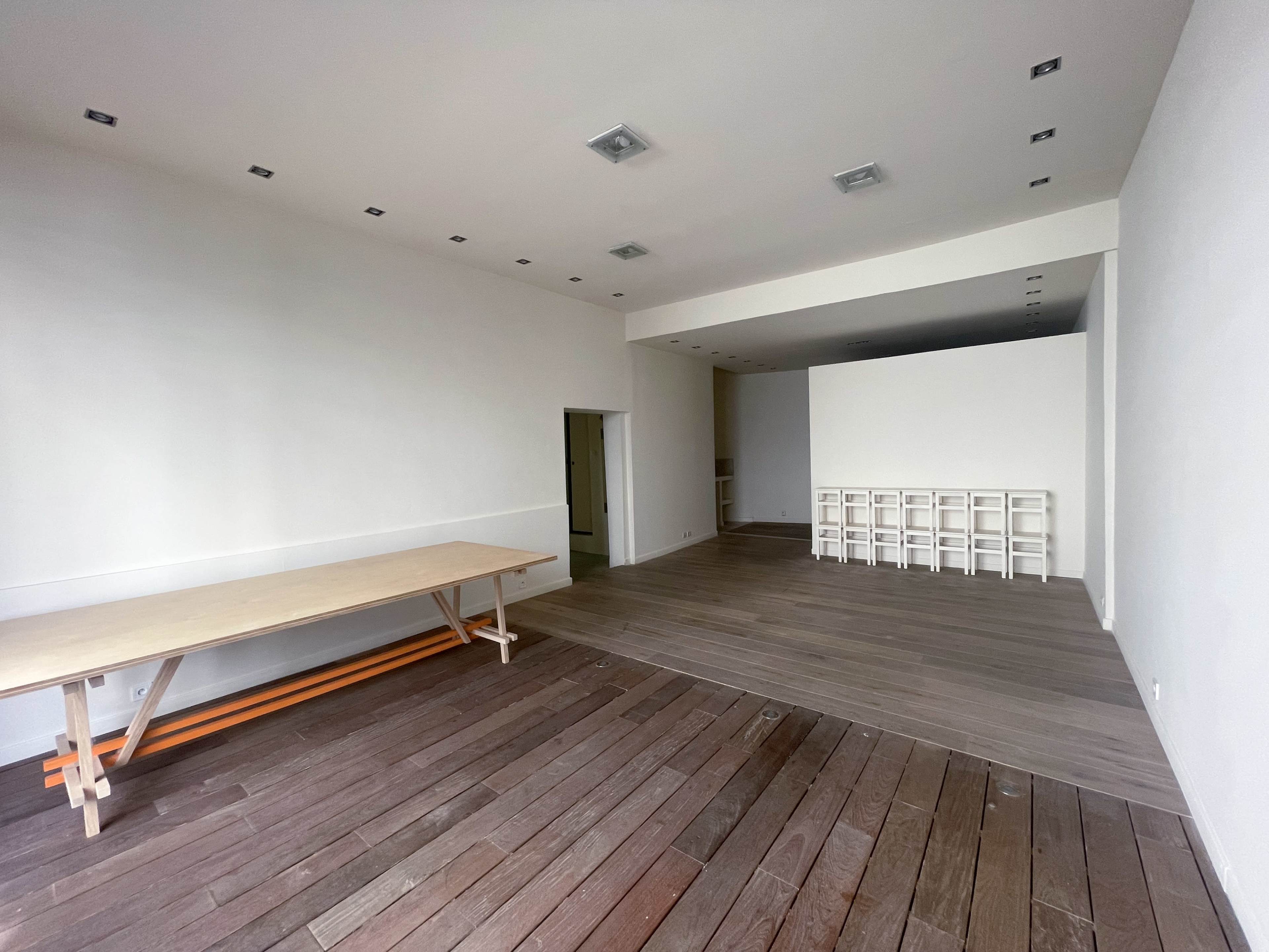 The image shows a spacious, empty room with wooden flooring, a long table on the left, and a section with white shelves against the far wall.