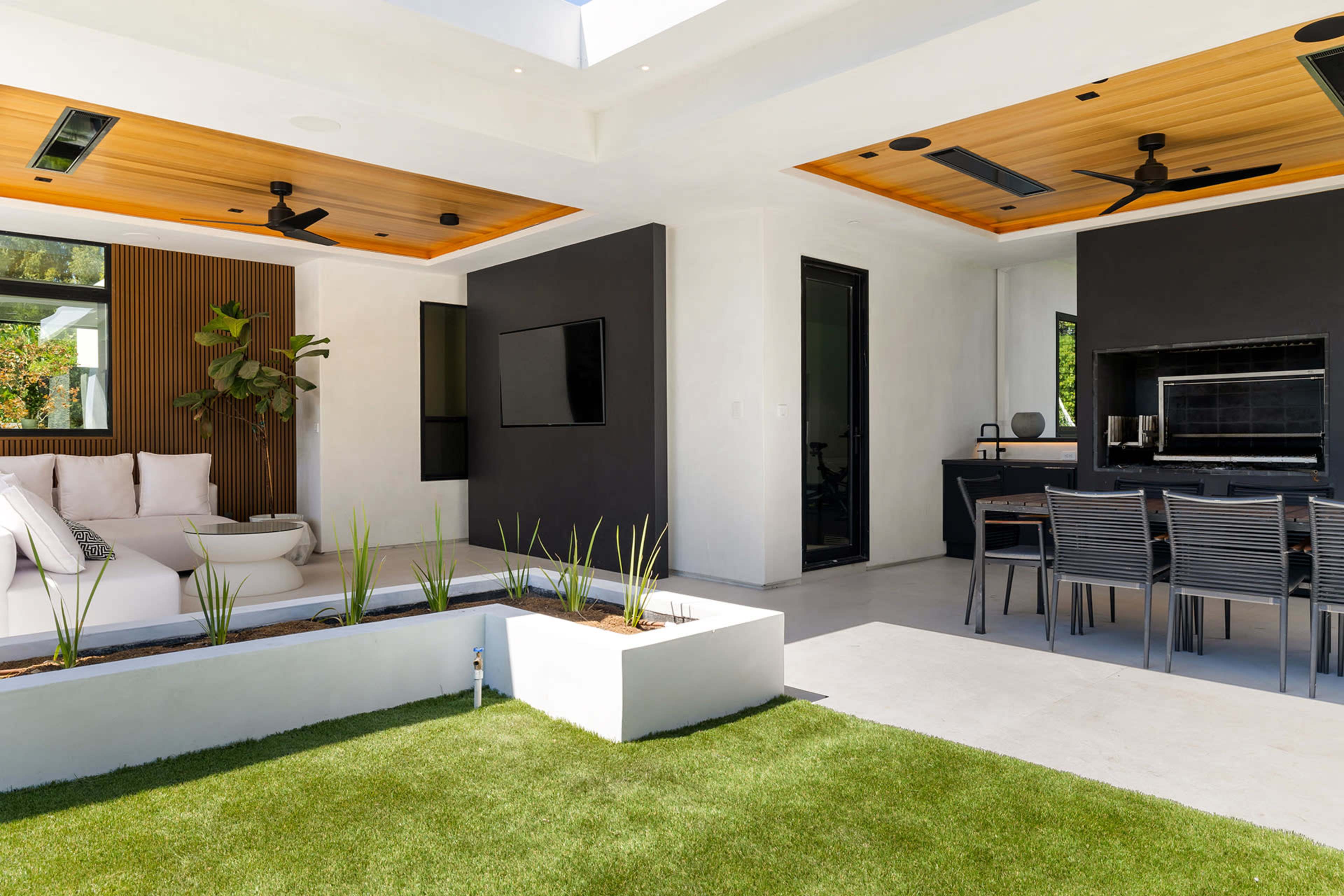 A modern outdoor living space features a seating area with a sofa and a dining table surrounded by greenery and a landscaped lawn.